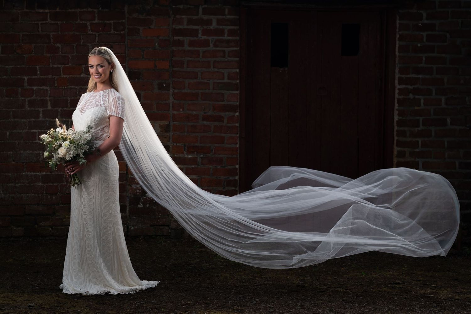 Bride in fitted white gown with dramatic cathedral veil flowing in wind against brick wall backdrop.