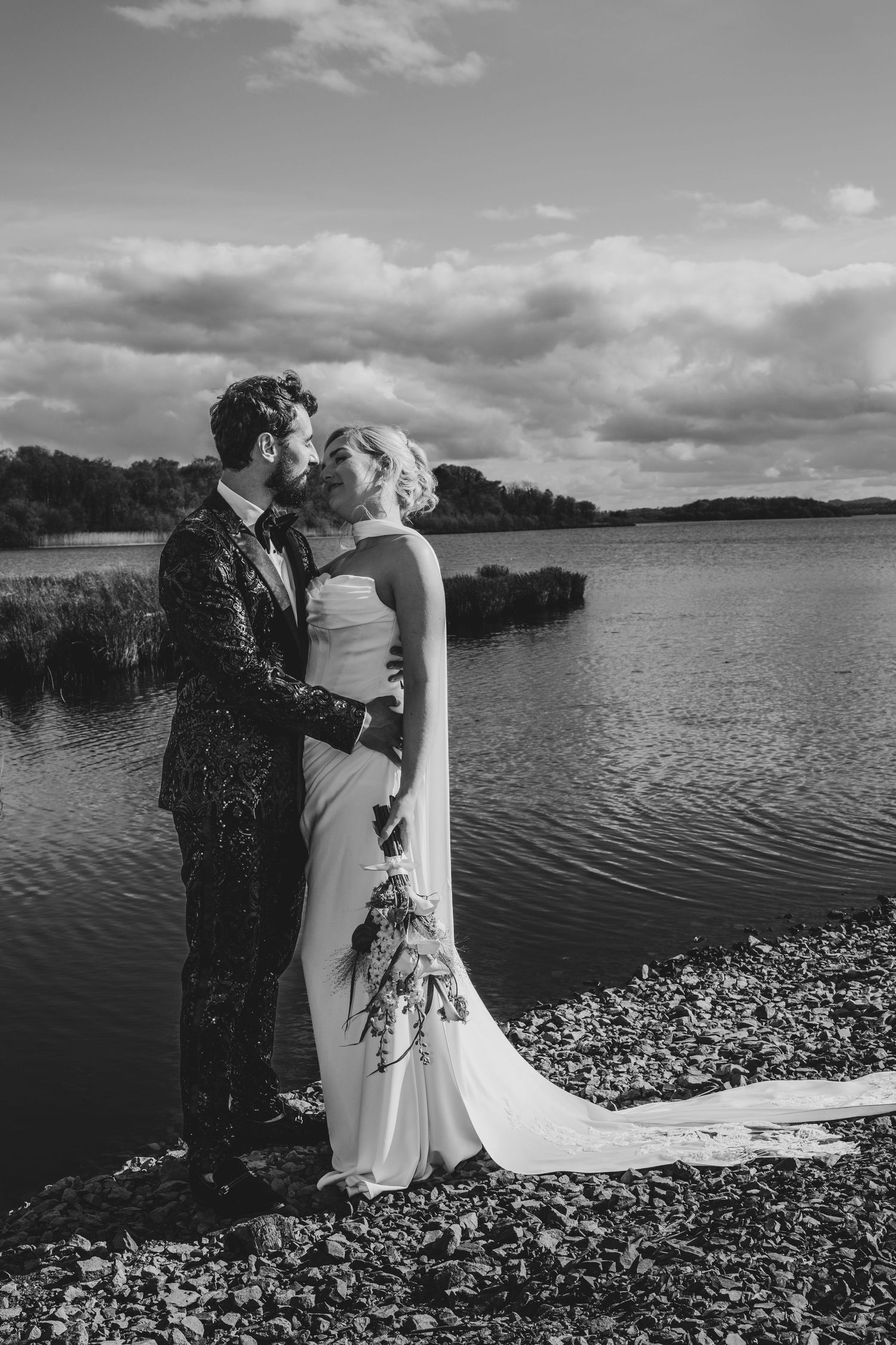 Bride and groom share kiss by scenic lake shore, groom in sequin jacket, bride in elegant white gown.