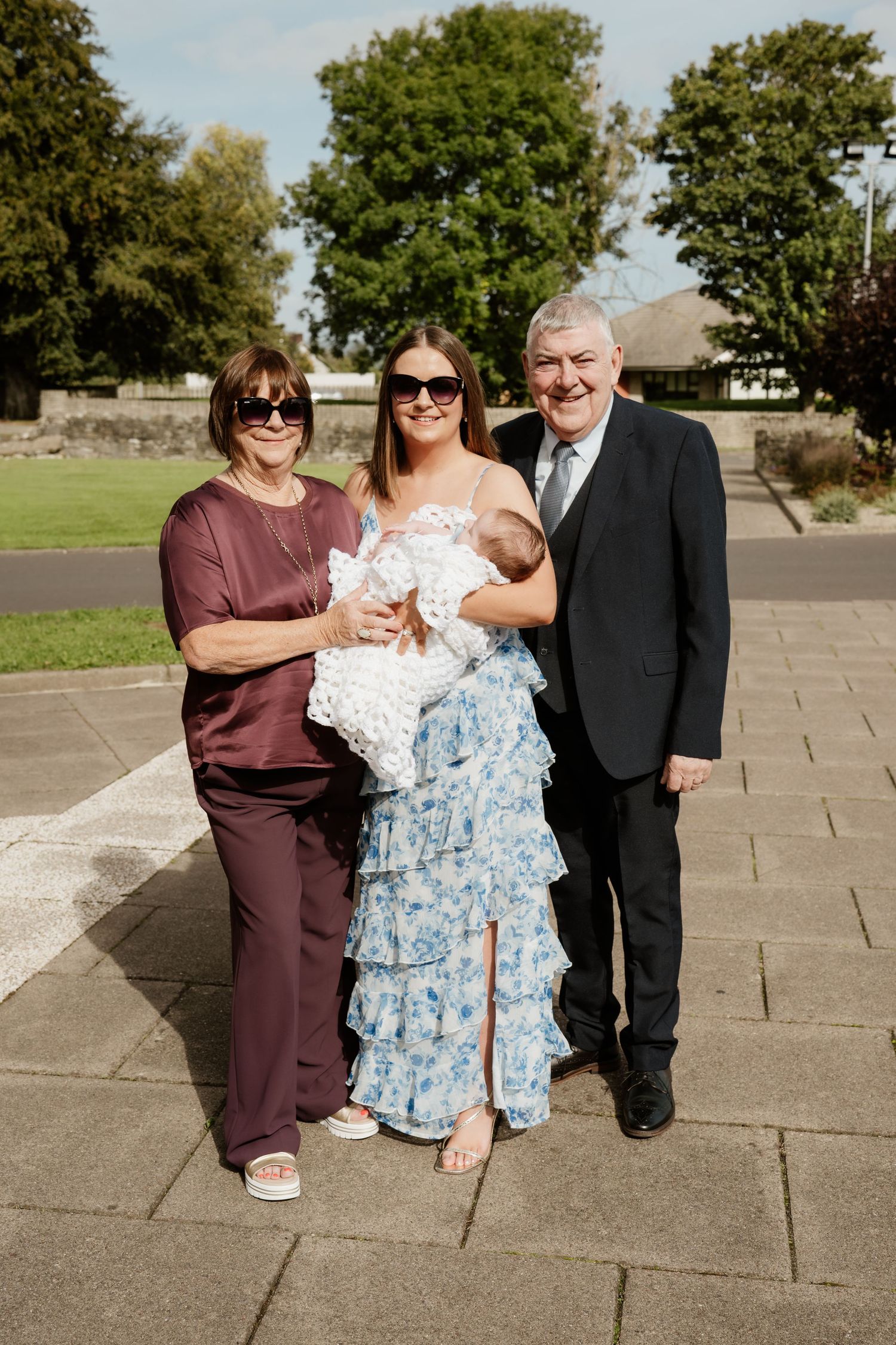 Family of three generations posing outdoors at a christening, with grandmother, mother holding baby, and grandfather.