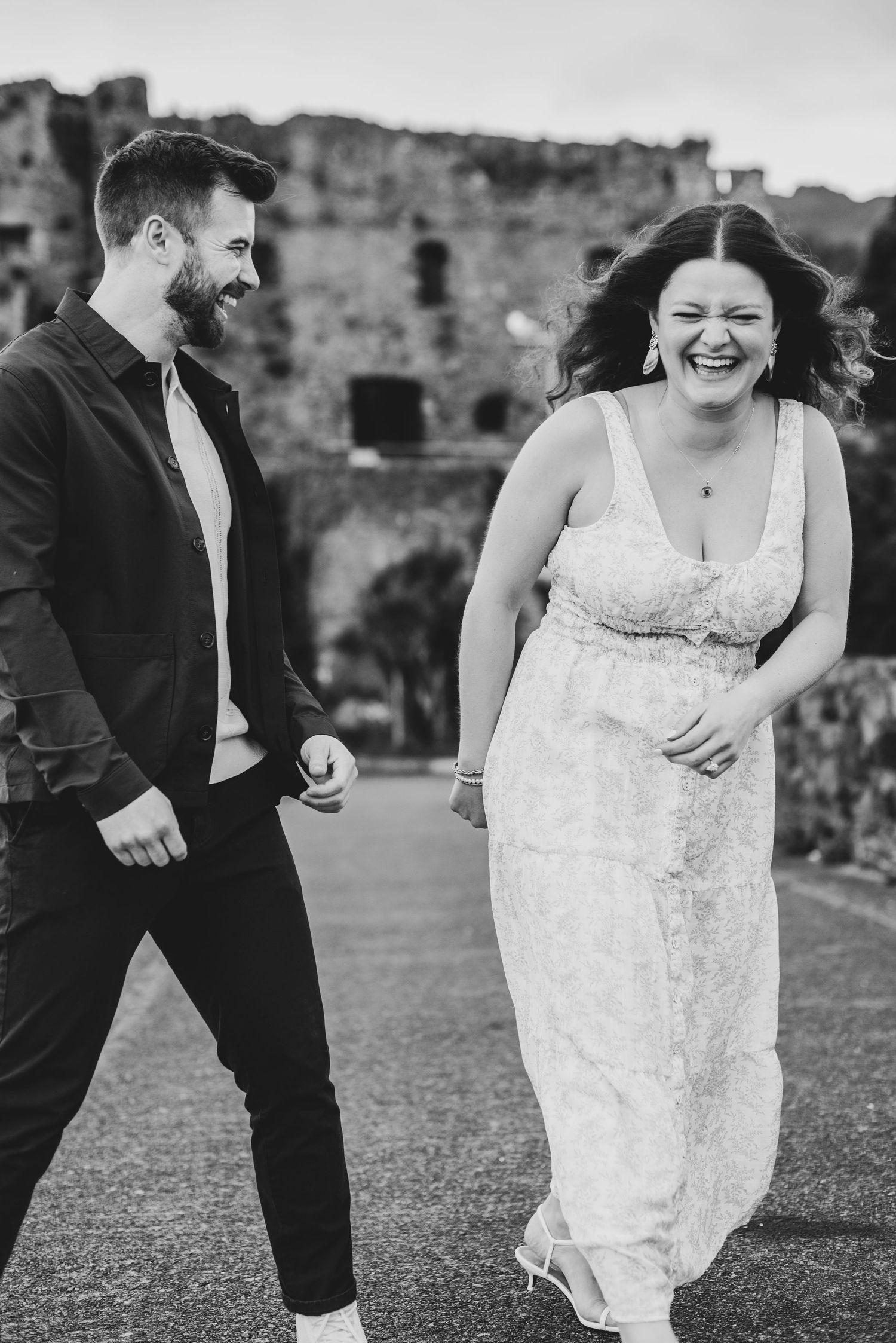Black and white photo of a laughing couple walking together outdoors near historic stone ruins.