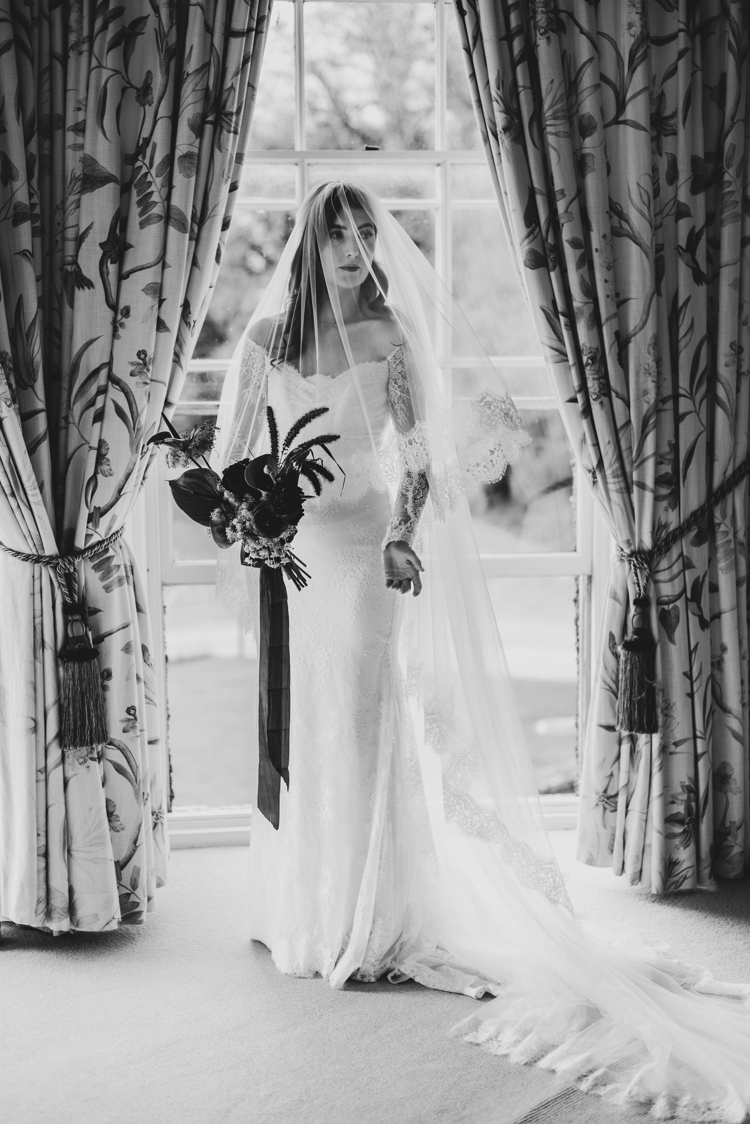 Bride in a fitted white gown and veil holds a dark bouquet, standing before a window with floral curtains.