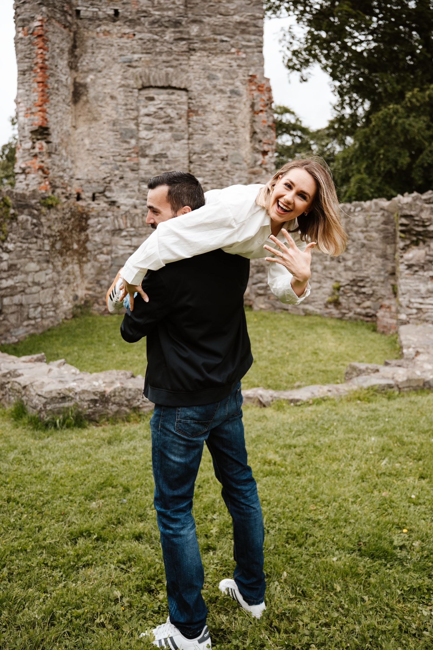 A joyful couple at castle ruins, man carrying woman on his back as she laughs and shows off her engagement ring.
