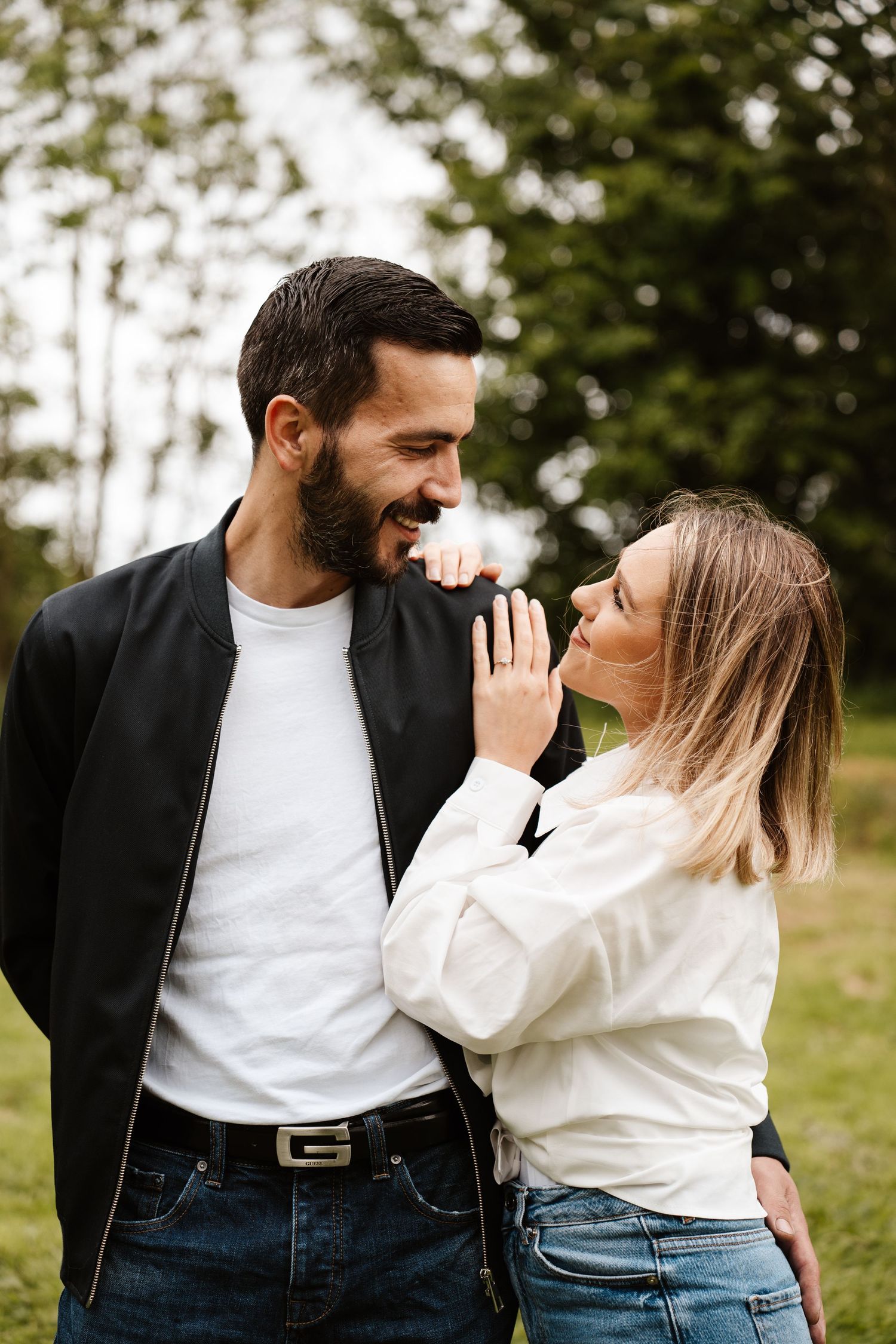 A smiling couple sharing a romantic moment outdoors, woman in white shirt touching man's face in a lush green park.
