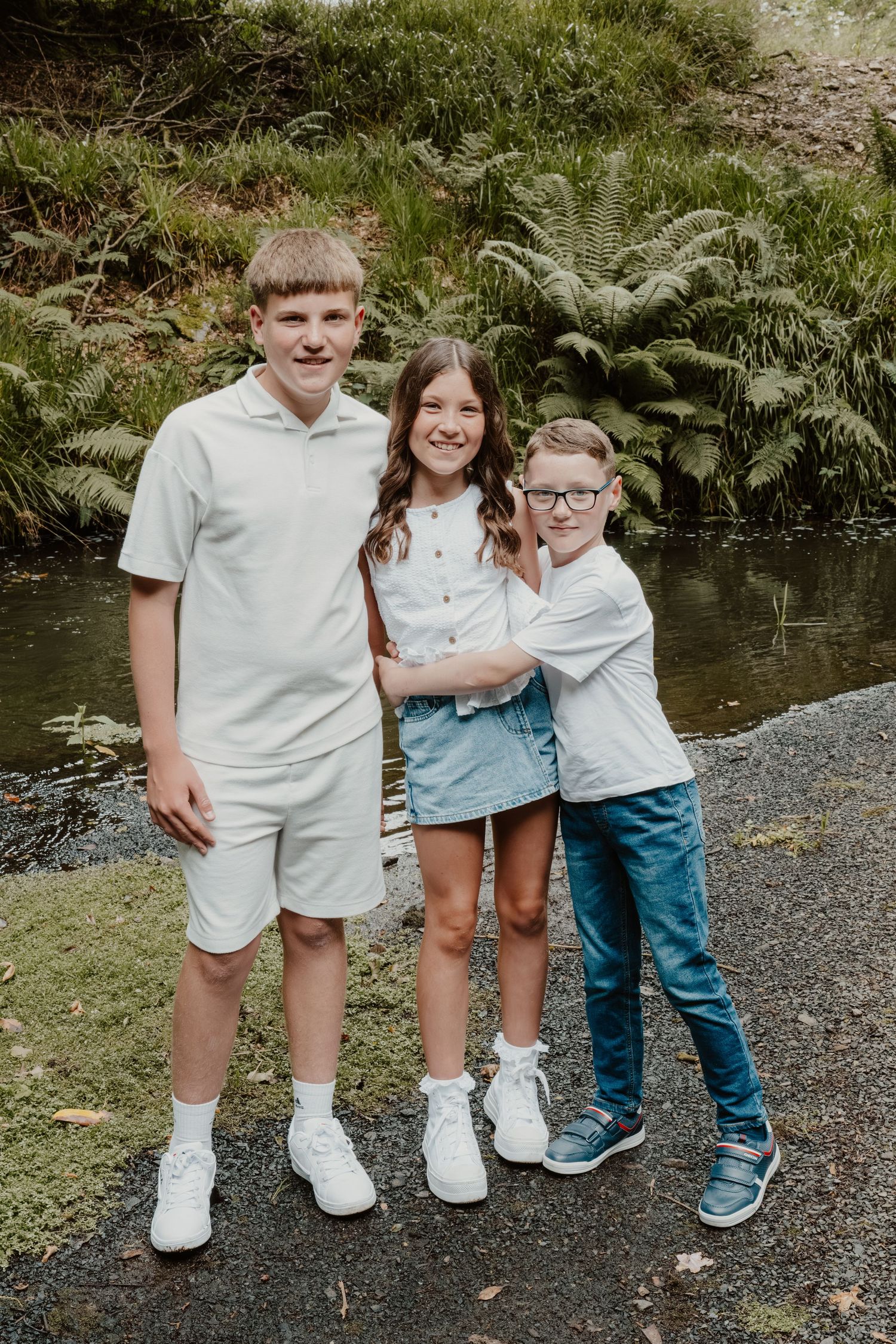 Three kids smiling and posing together outdoors near a creek surrounded by lush green ferns.
