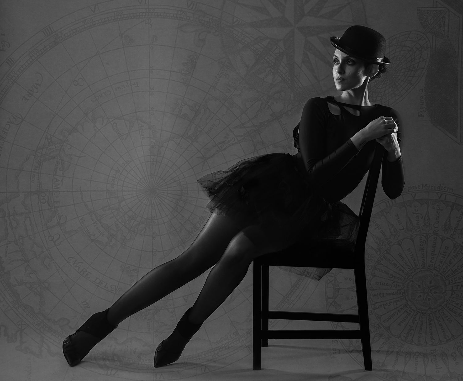 Black and white photo of a ballerina in tutu and bowler hat seated gracefully on a chair.