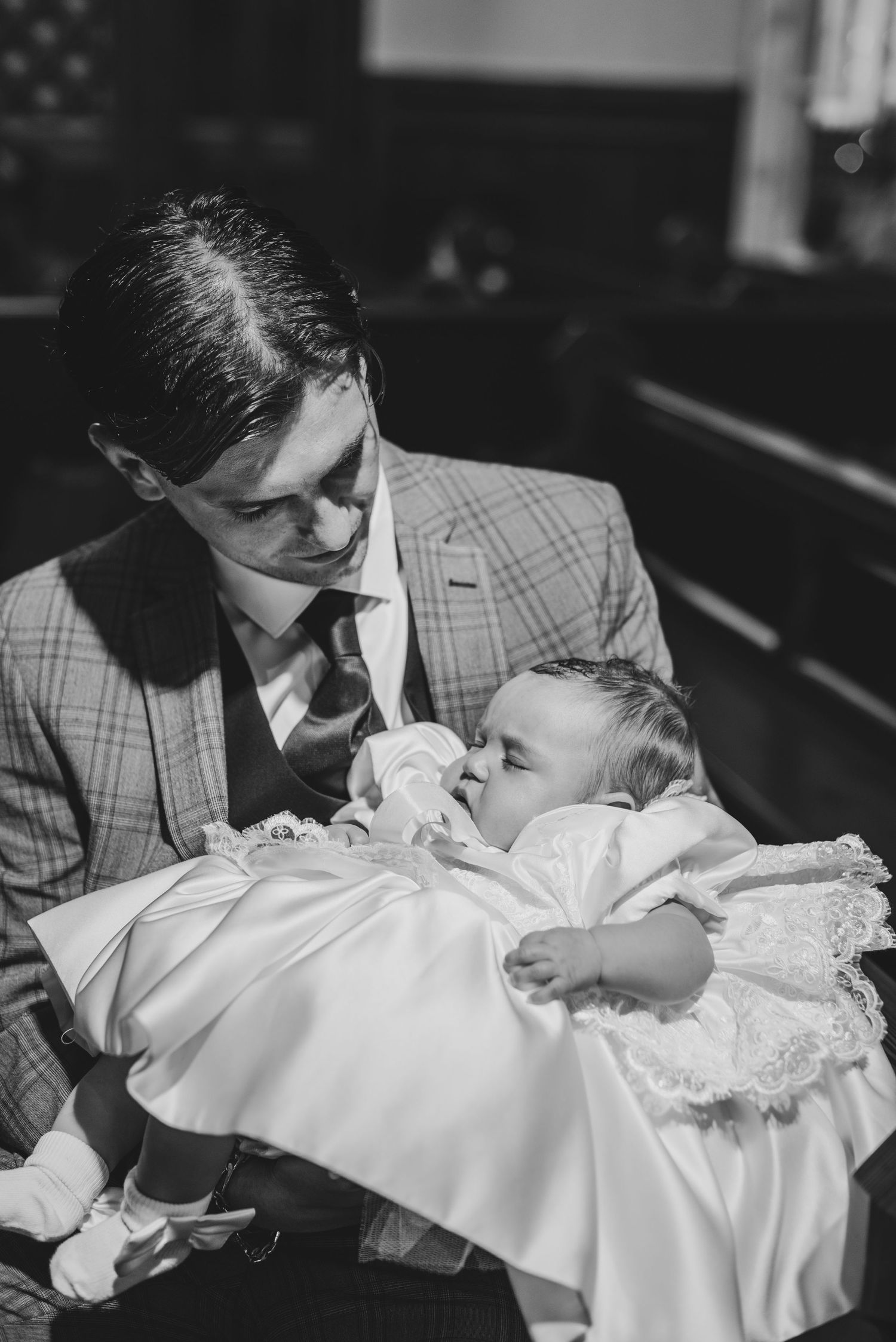 Black and white photo of a man in a plaid suit holding a baby in a white christening gown inside a church.