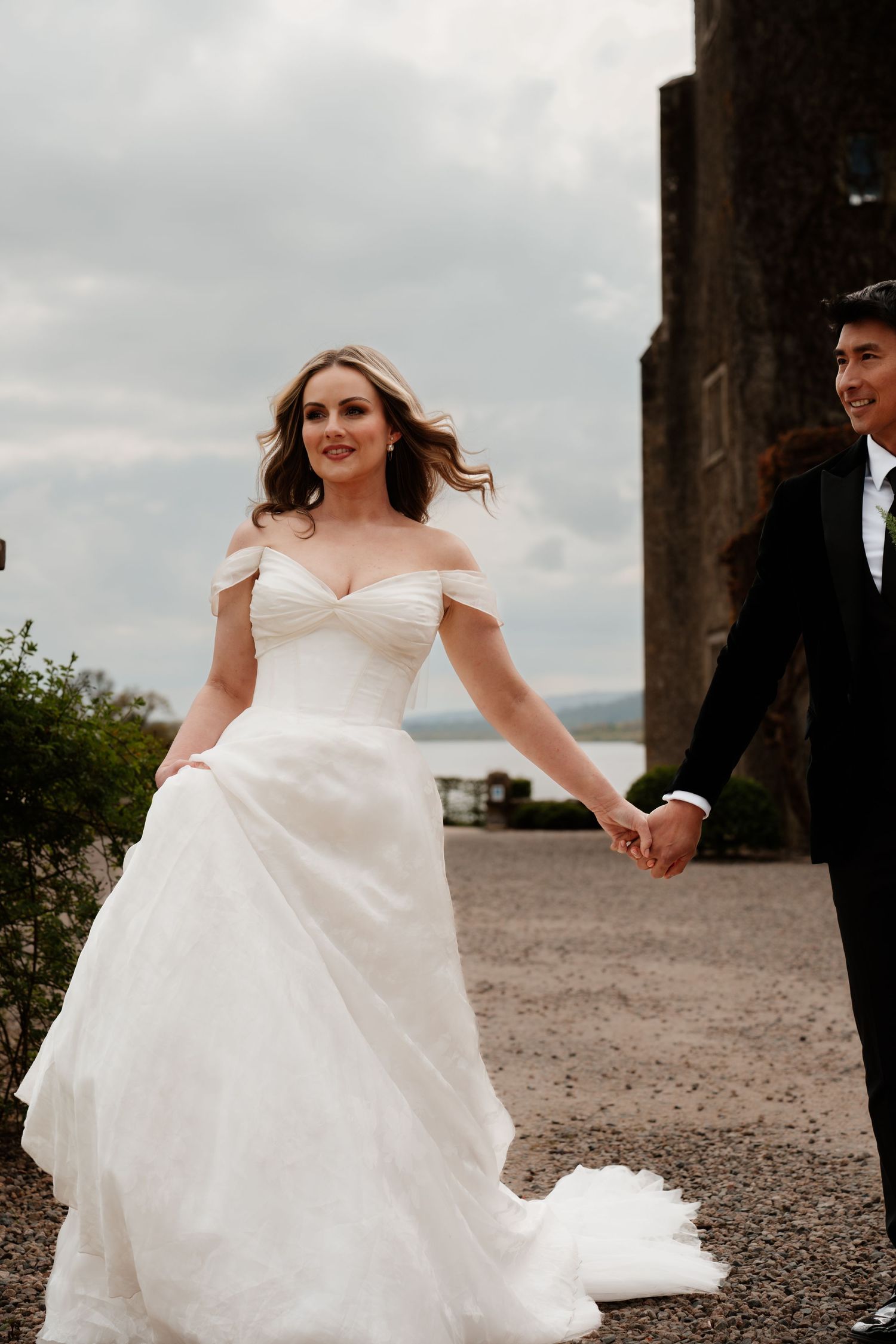 Bride in off-shoulder white gown holds groom's hand near stone castle under cloudy sky.