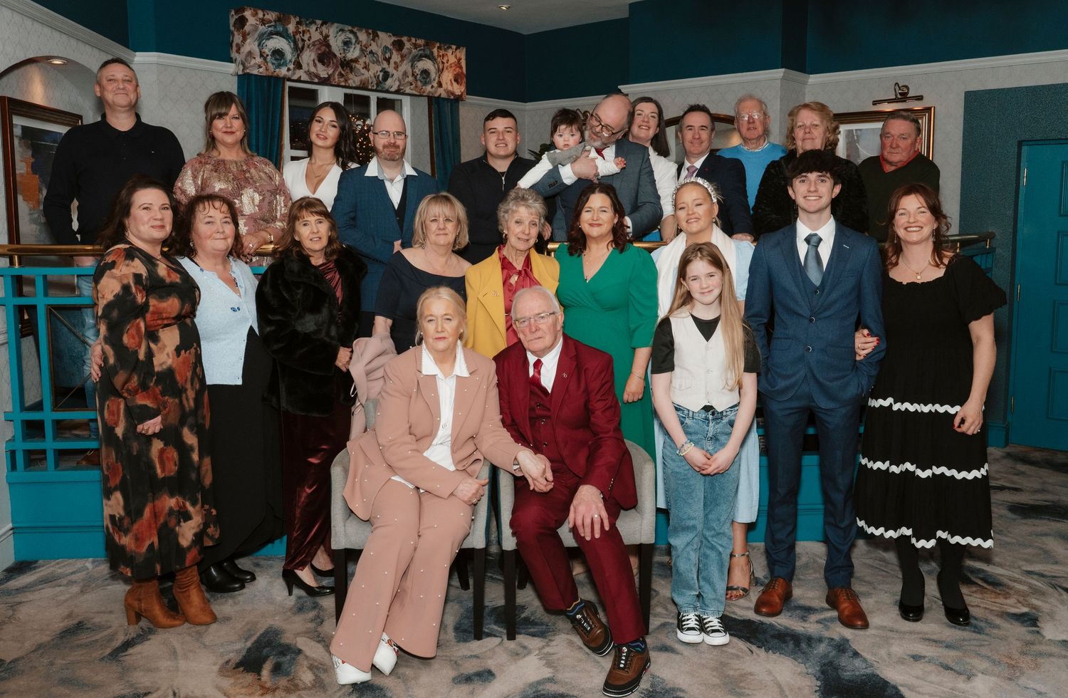 Large group of formally dressed adults posing together for a group photo in a decorated indoor setting.