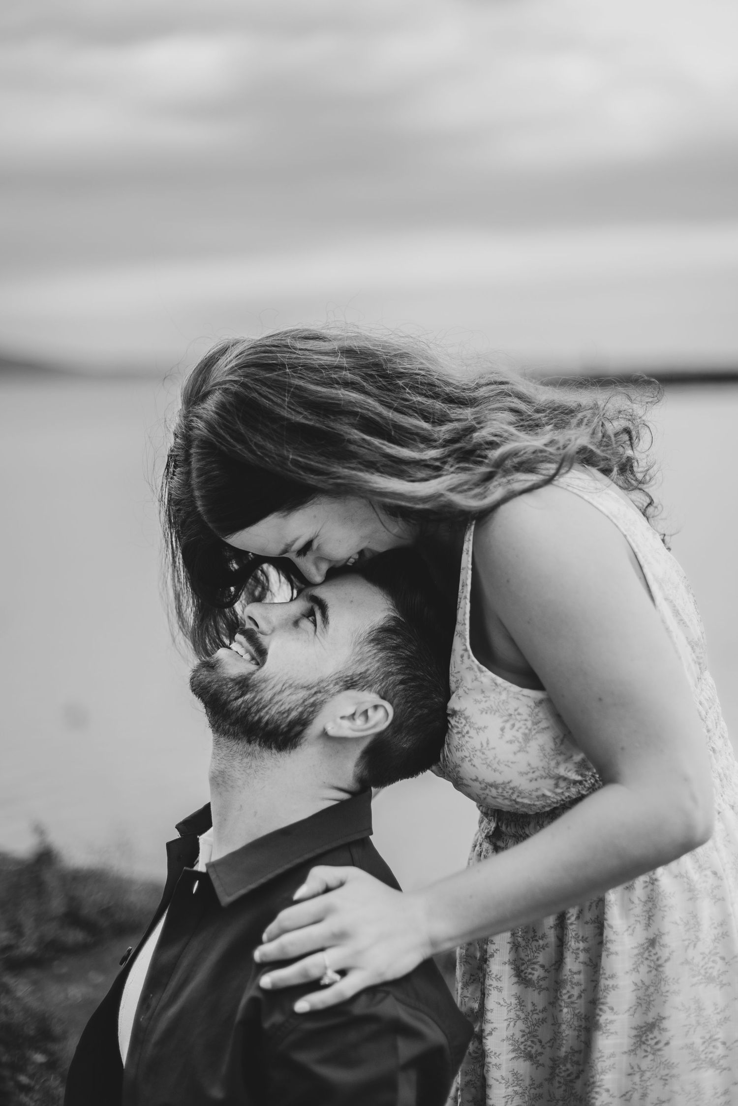 Black and white romantic photo of couple about to kiss outdoors near water with cloudy sky.