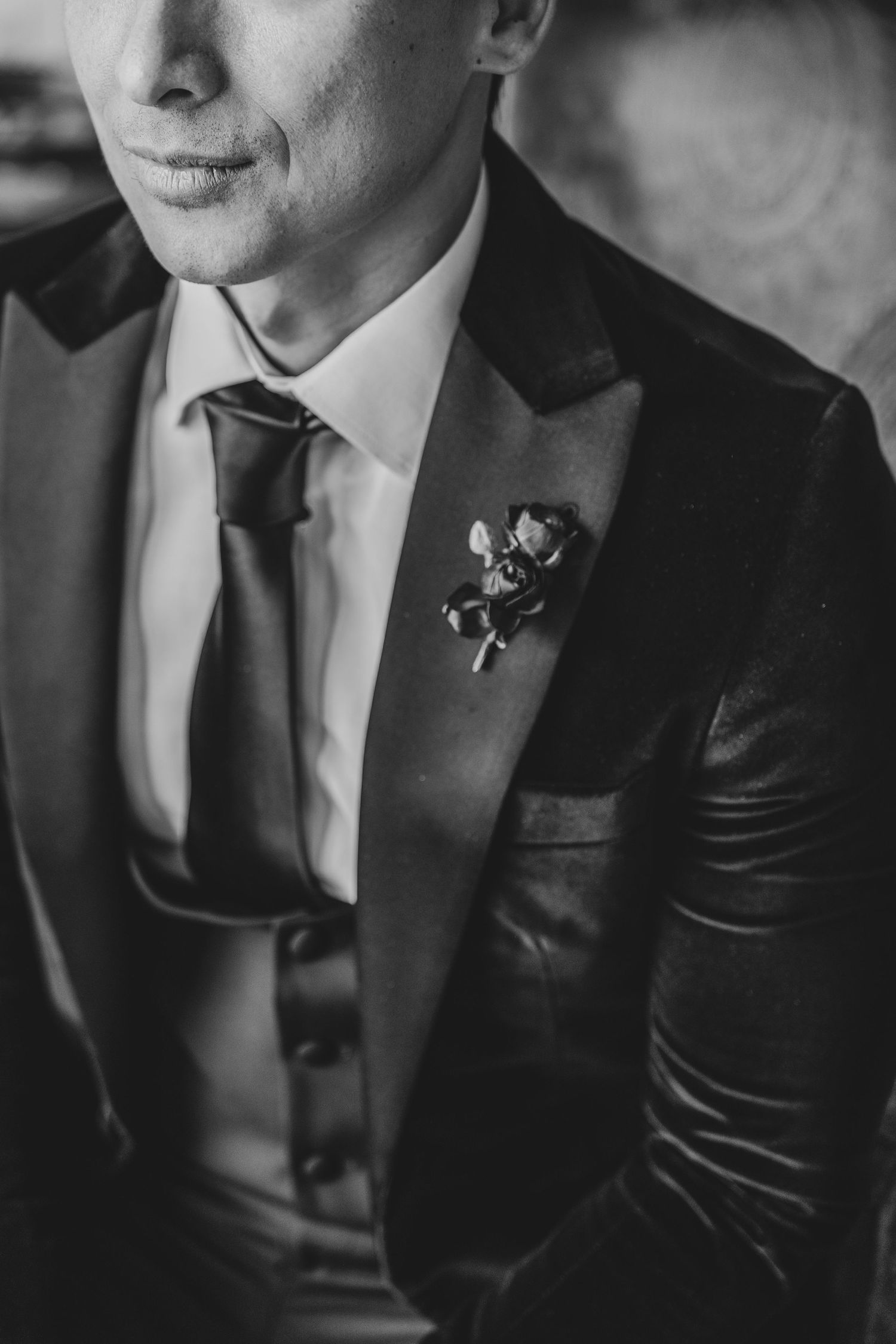 Black and white close-up of groom in velvet tuxedo with boutonniere and satin tie.
