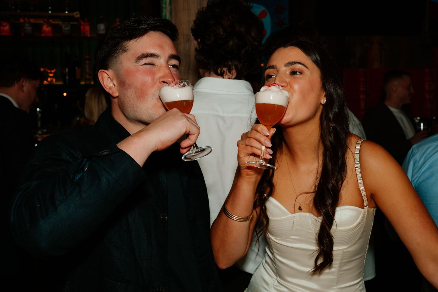 Bride and groom drinking Belgian beer glasses together at wedding reception party.