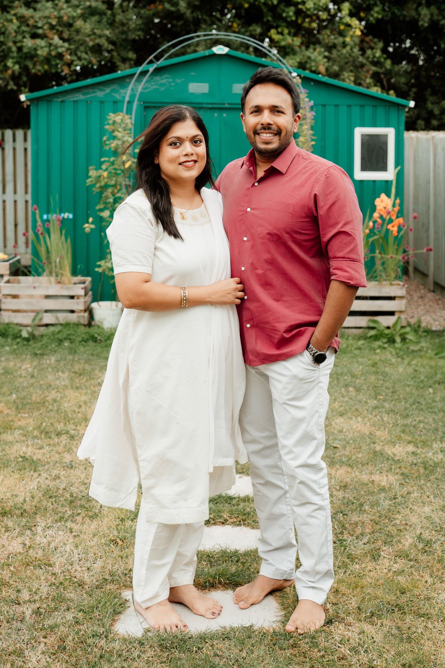 A smiling couple stands barefoot in a backyard, woman in white kurta, man in red shirt and white pants.