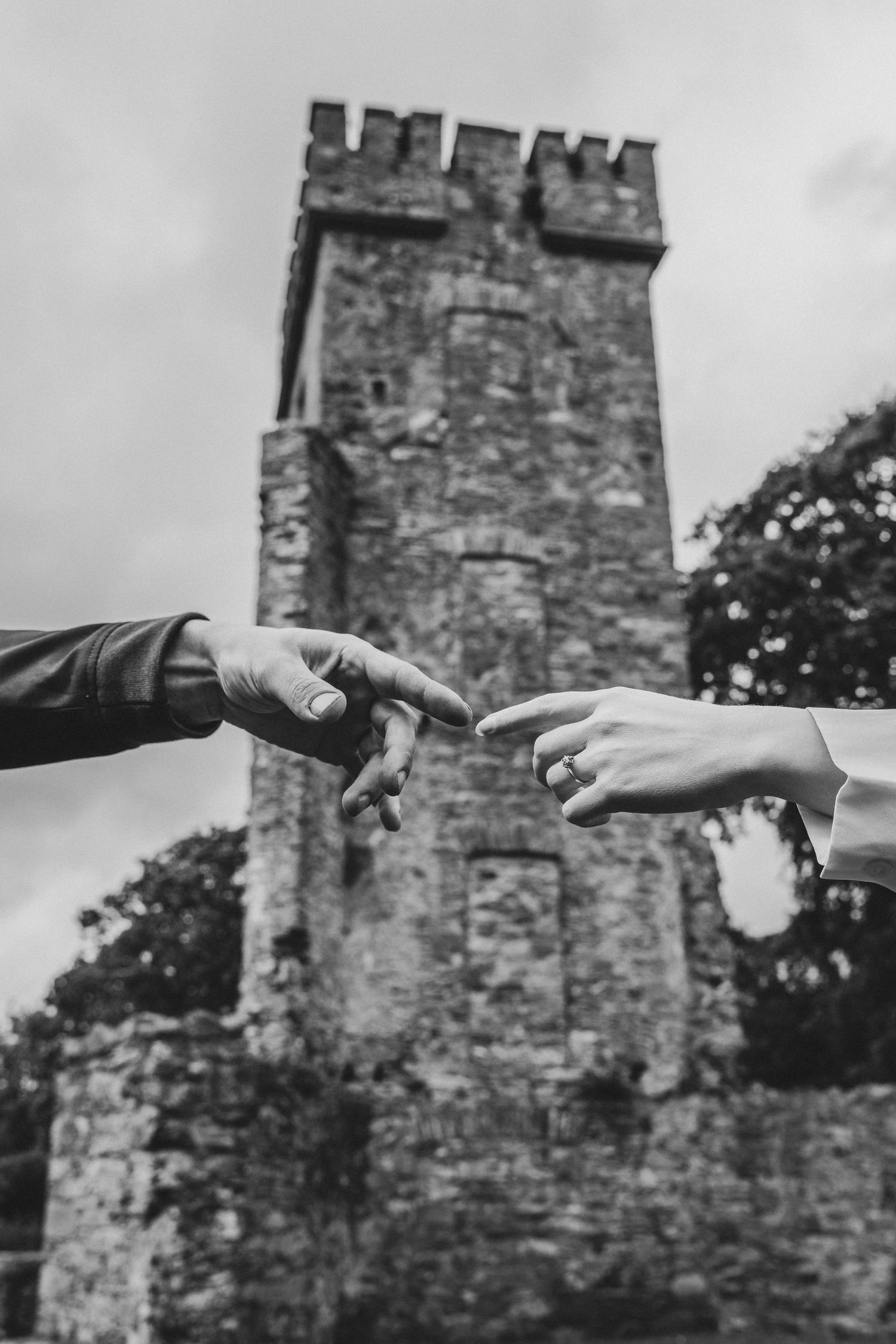 Black and white photo of two hands nearly touching in front of a historic stone castle tower, evoking romance.