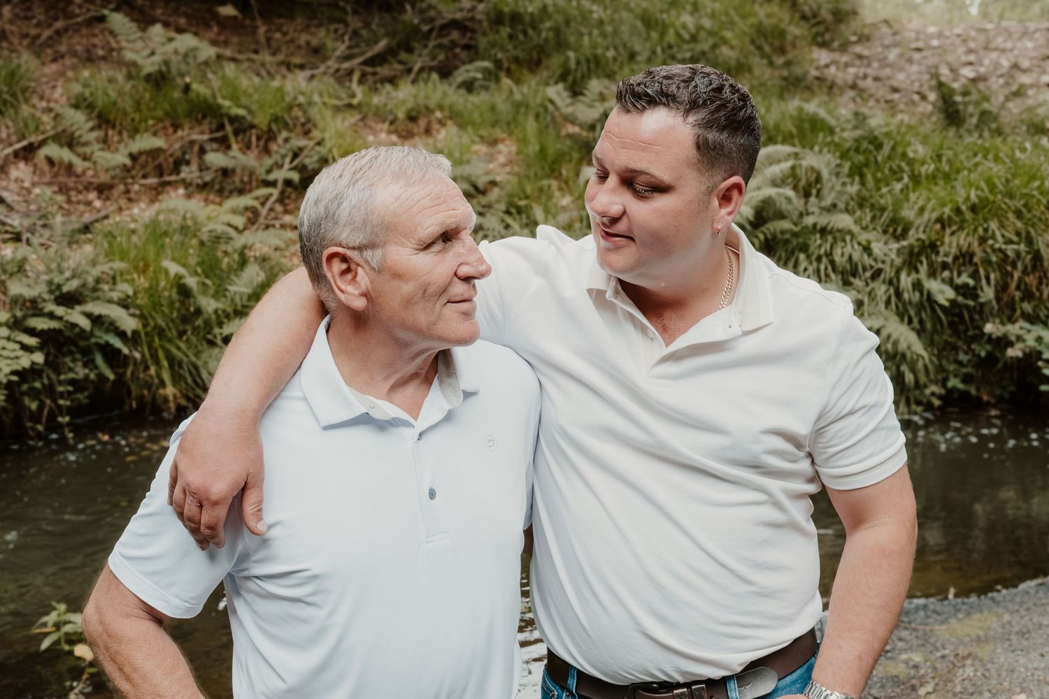 Two men smiling and embracing outdoors near a stream with lush green foliage in the background.
