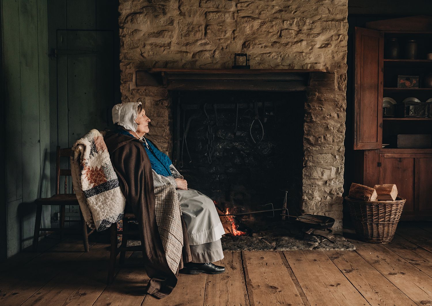 Elderly woman sitting in rocking chair by stone fireplace on wooden floor, wrapped in quilt blanket.