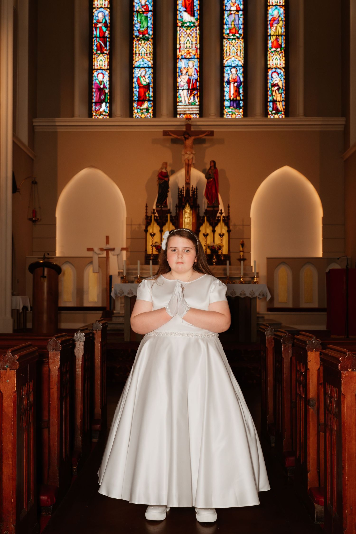 Young girl in white dress praying at church altar during First Communion ceremony with stained glass windows.