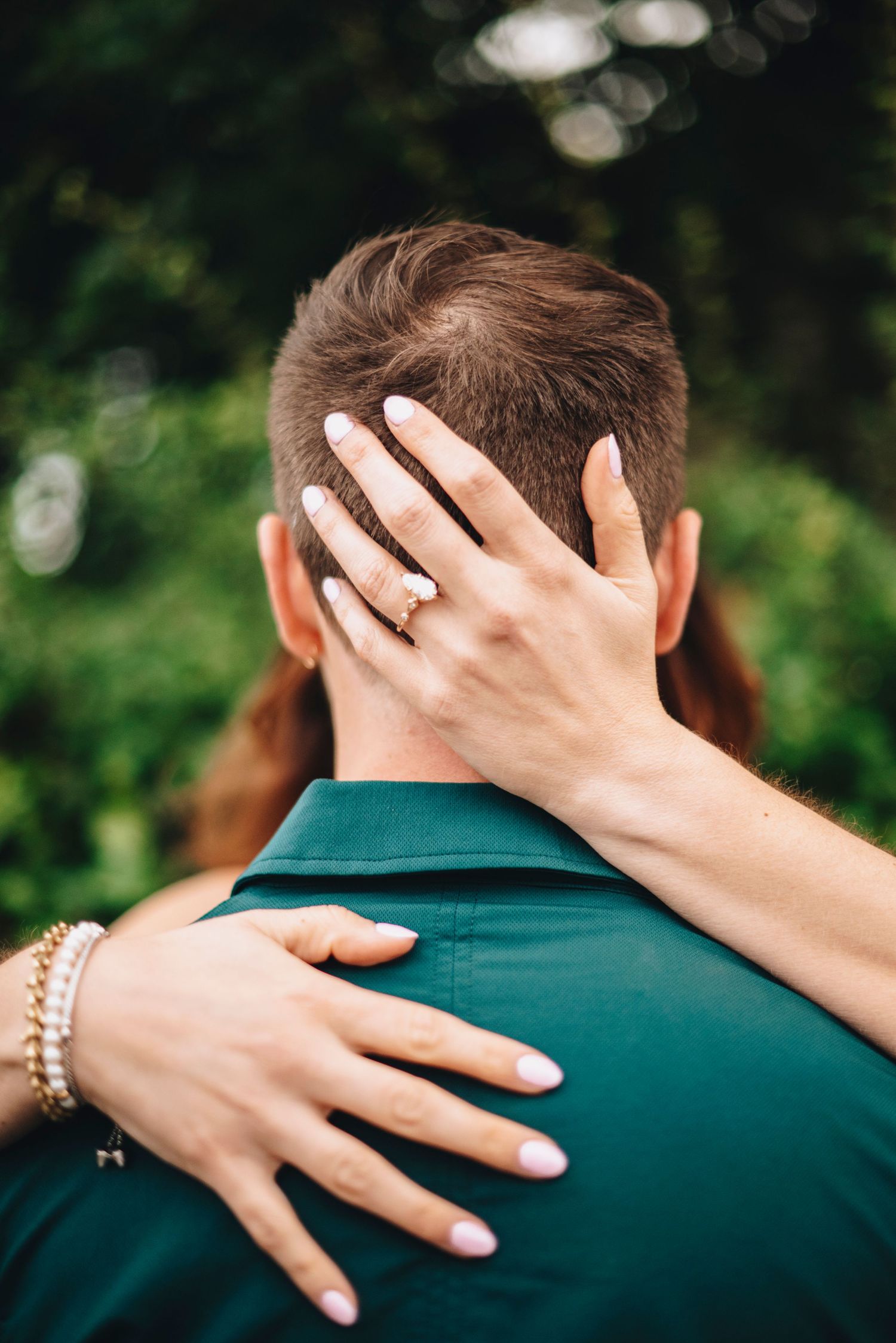 Close-up of woman's hand with gold engagement ring resting on back of man's head in green shirt.