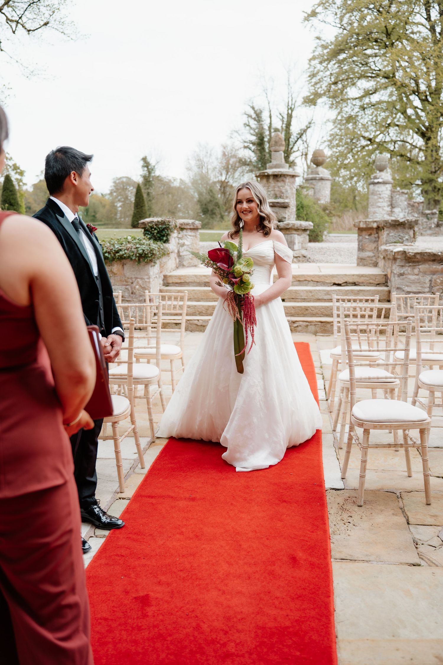 Smiling bride in a white ball gown walks down a red carpet aisle at an outdoor garden wedding ceremony.