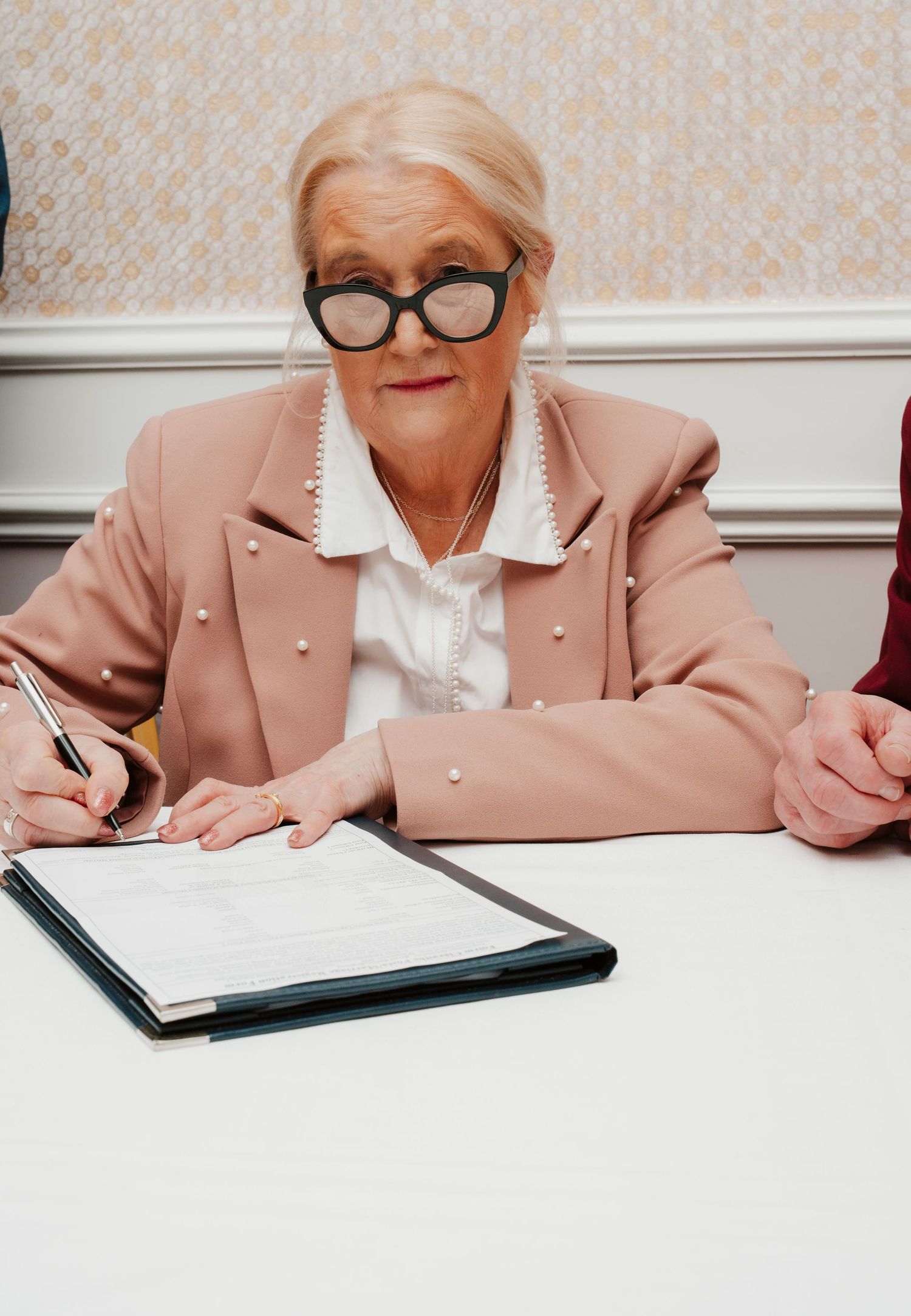 Elderly woman in pink blazer and black glasses signing documents at a white table.