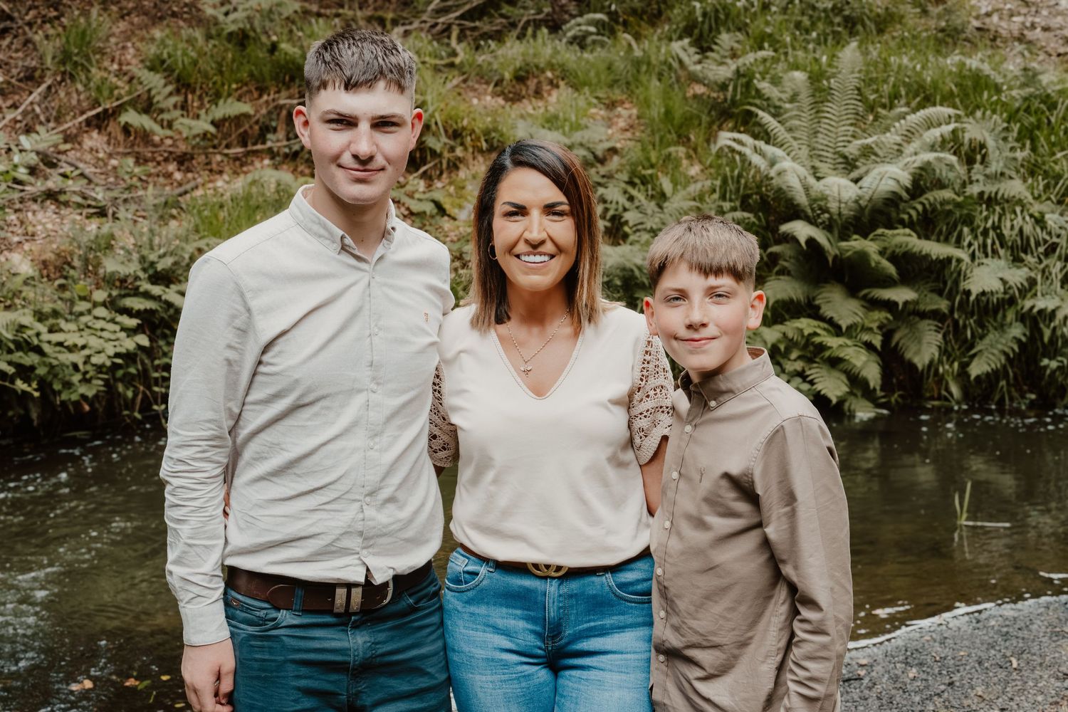 A woman and two boys smile together outdoors near a stream surrounded by lush green ferns.