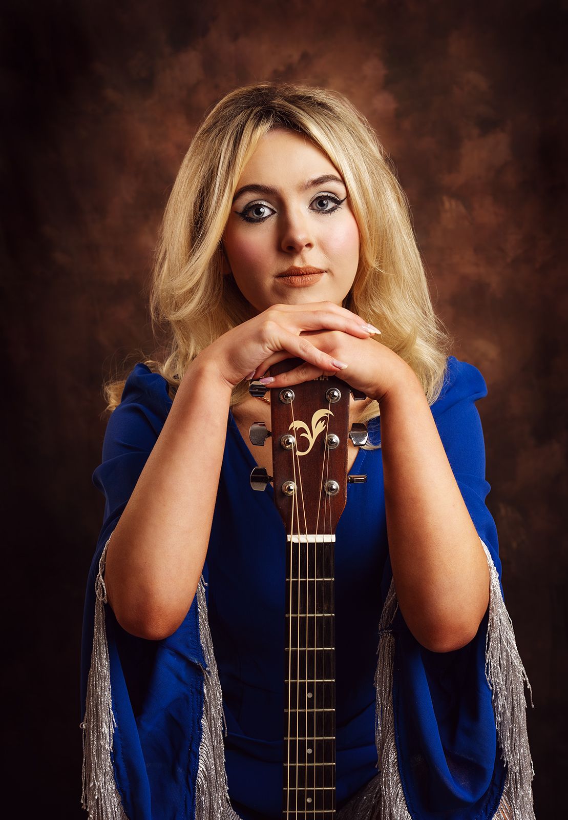 Young blonde woman in blue dress posing with acoustic guitar in professional studio portrait photography.