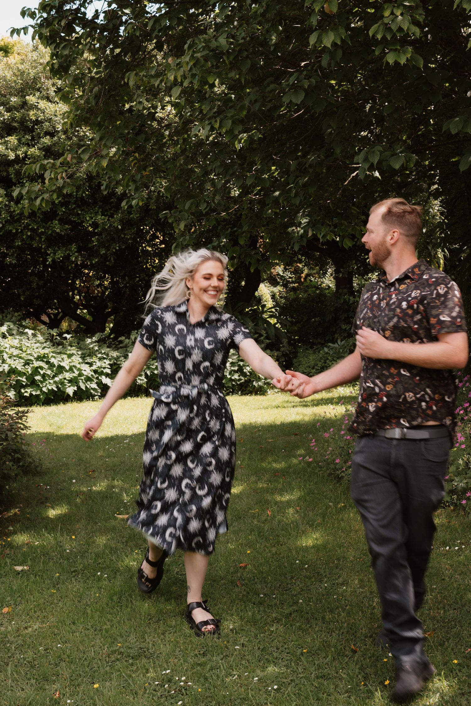 A joyful couple holds hands while walking through a lush green garden, the woman wearing a floral dress and smiling.