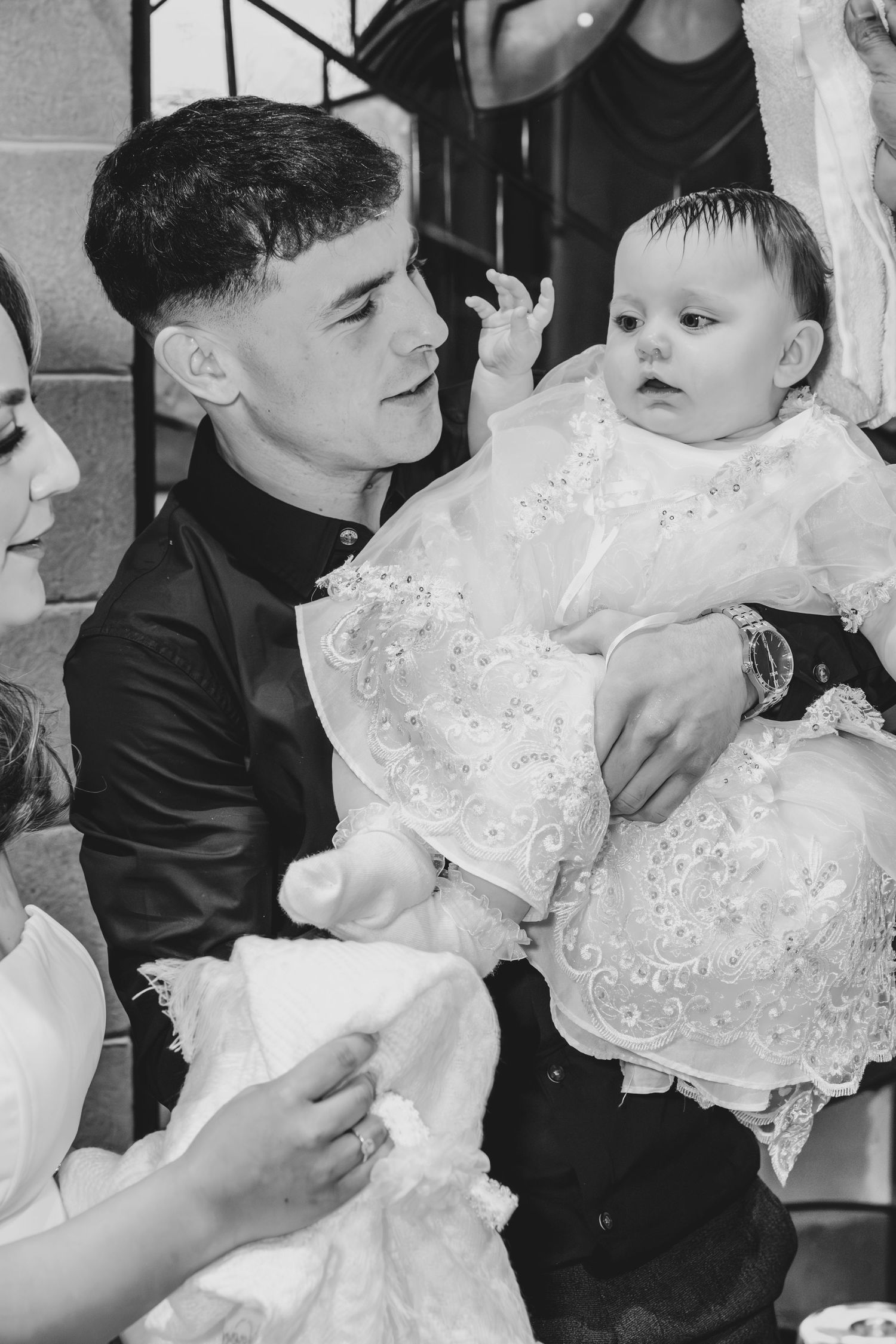 Black and white photo of a young man holding a baby in a white christening gown at a baptism ceremony.
