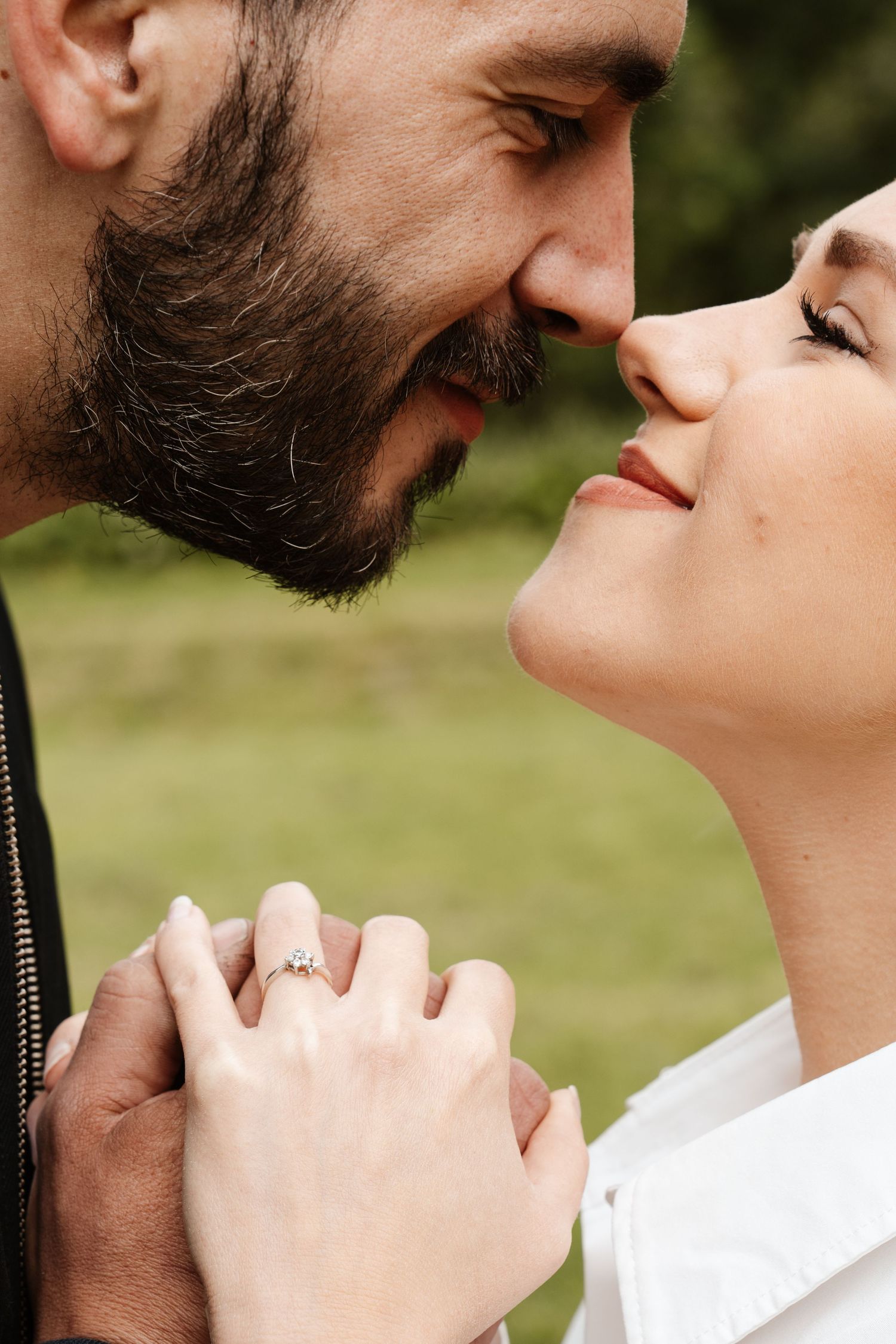 Couple almost kissing while holding hands showing an engagement ring, with a green park background.