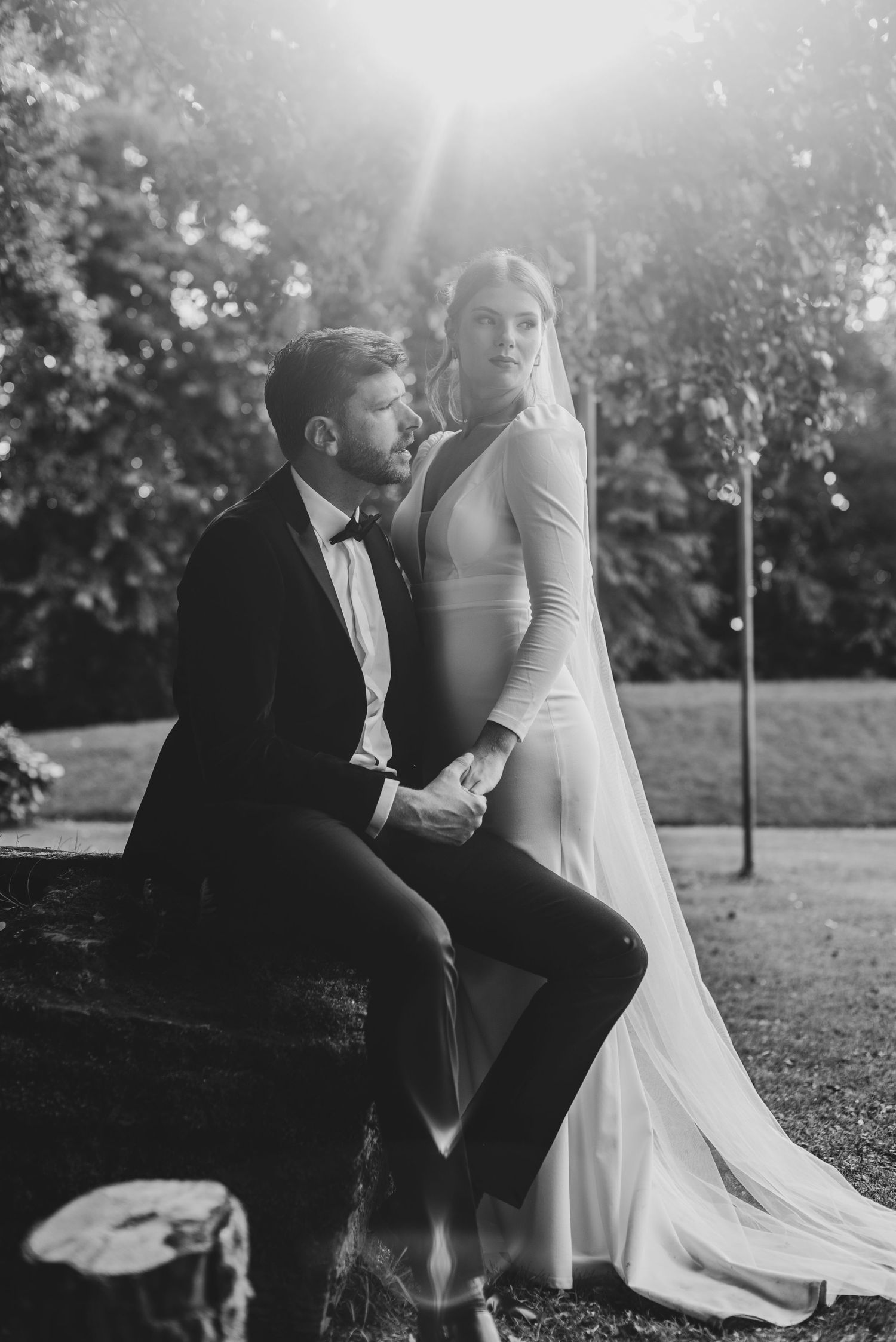 Black and white romantic portrait of bride and groom sitting outdoors with sunlight glowing behind trees.