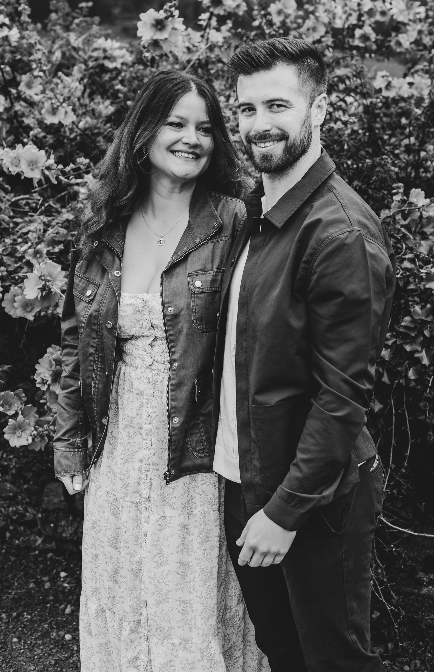 Couple smiling together outdoors in leather jackets against a floral background in black and white.