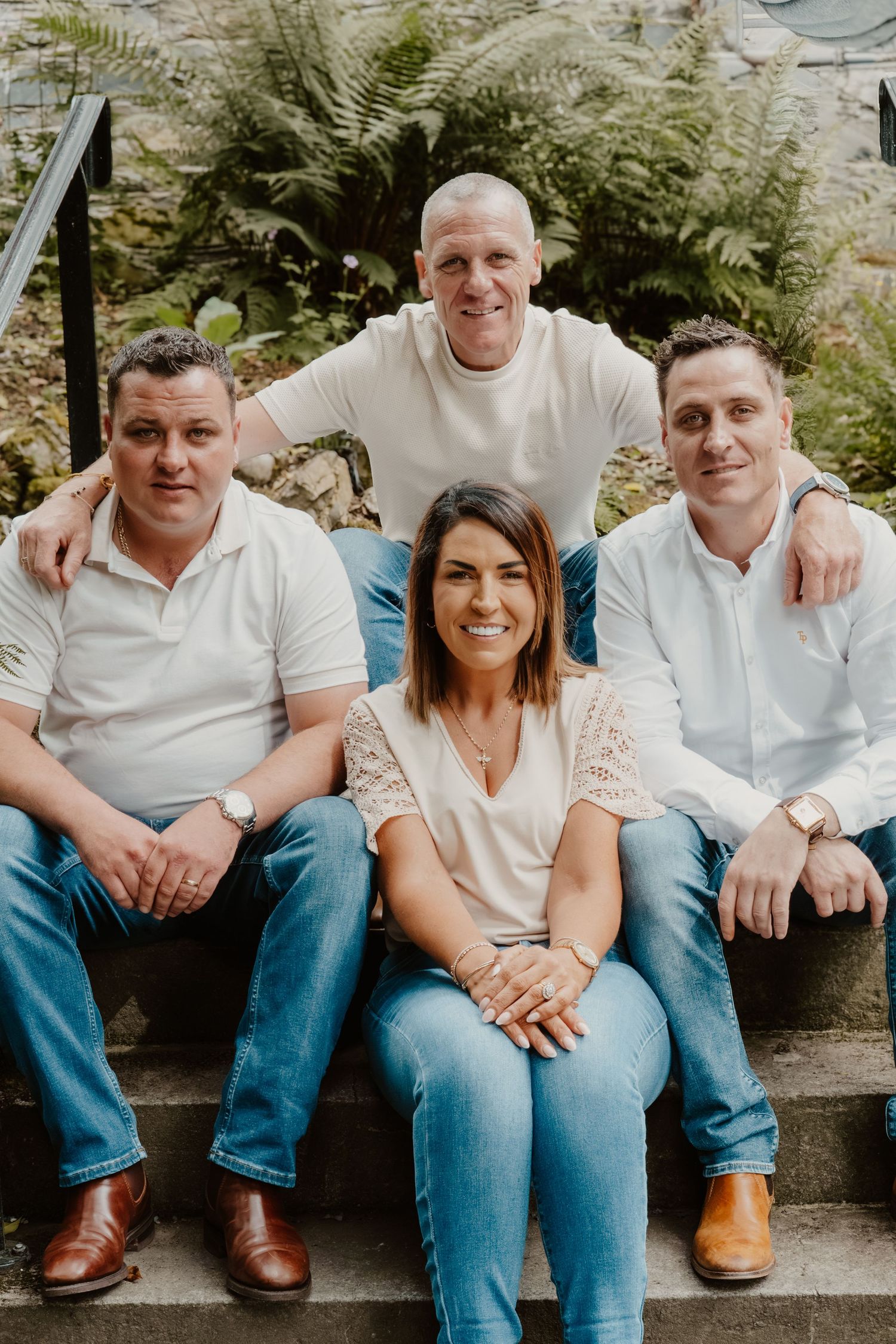 Four smiling adults pose on stone steps surrounded by lush green ferns in a casual outdoor group portrait.