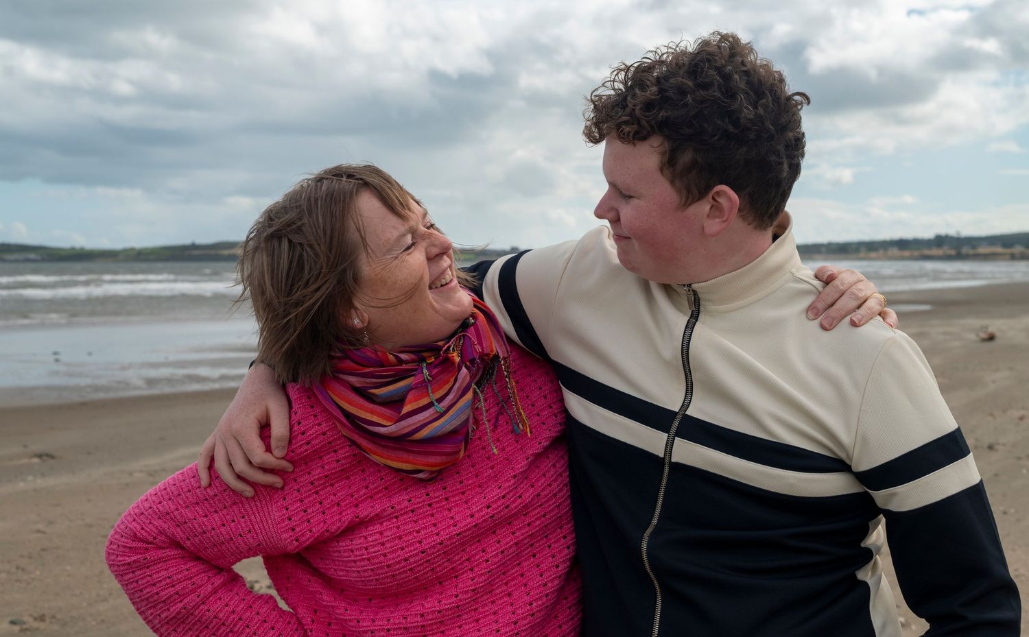 Two people smiling and embracing on a sandy beach with cloudy skies, wearing casual colorful clothing.