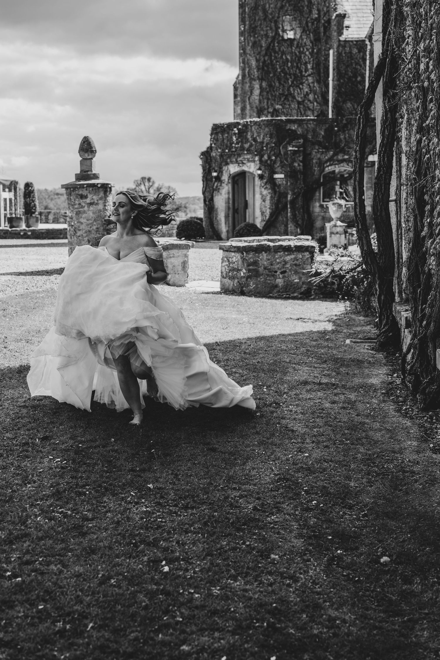 Black and white photo of bride in flowing gown running beside historic stone castle exterior.