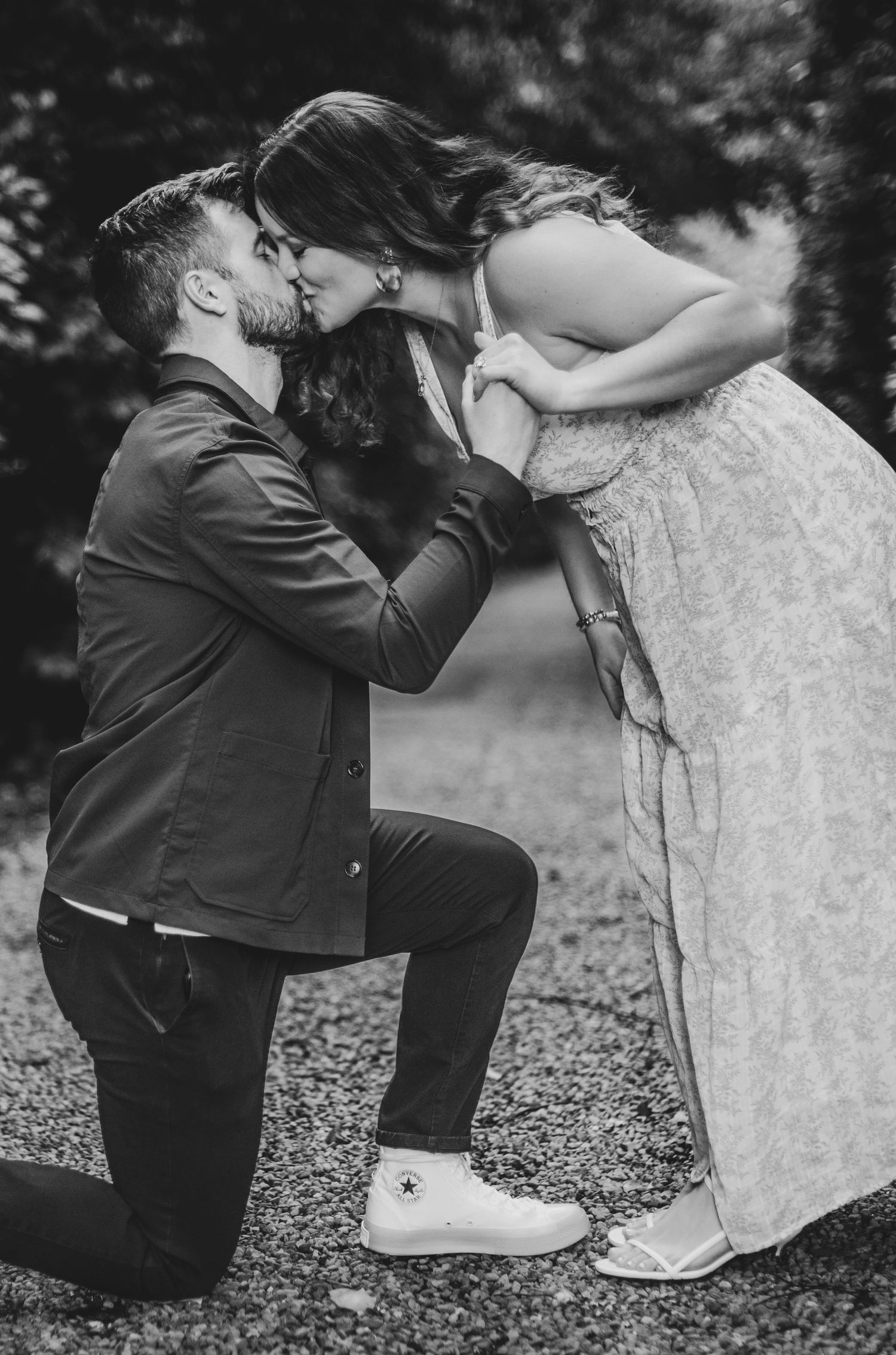 Black and white photo of a man on one knee kissing his partner during a romantic outdoor engagement moment.