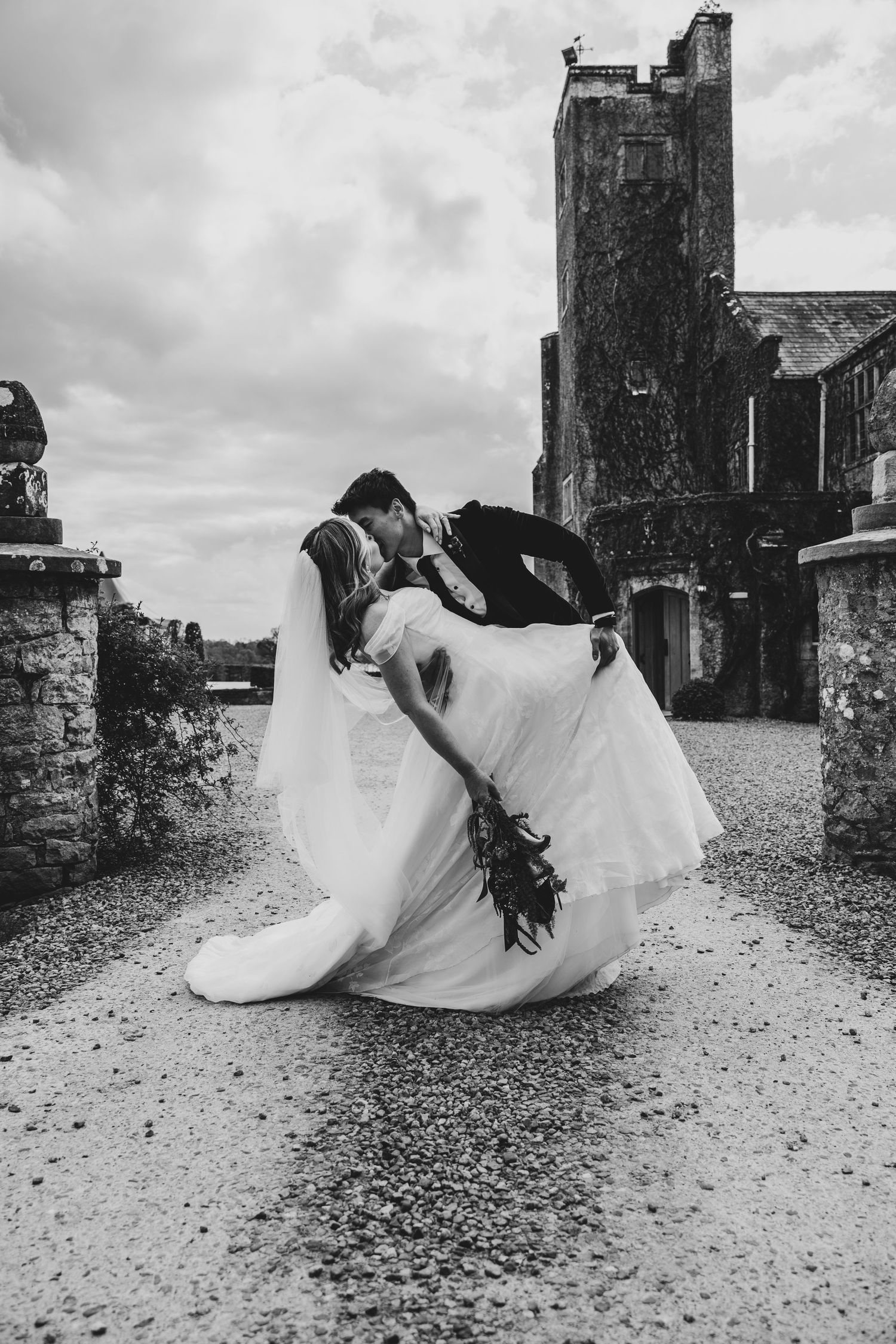 Black and white photo of a groom dipping his bride for a kiss outside a historic castle on their wedding day.