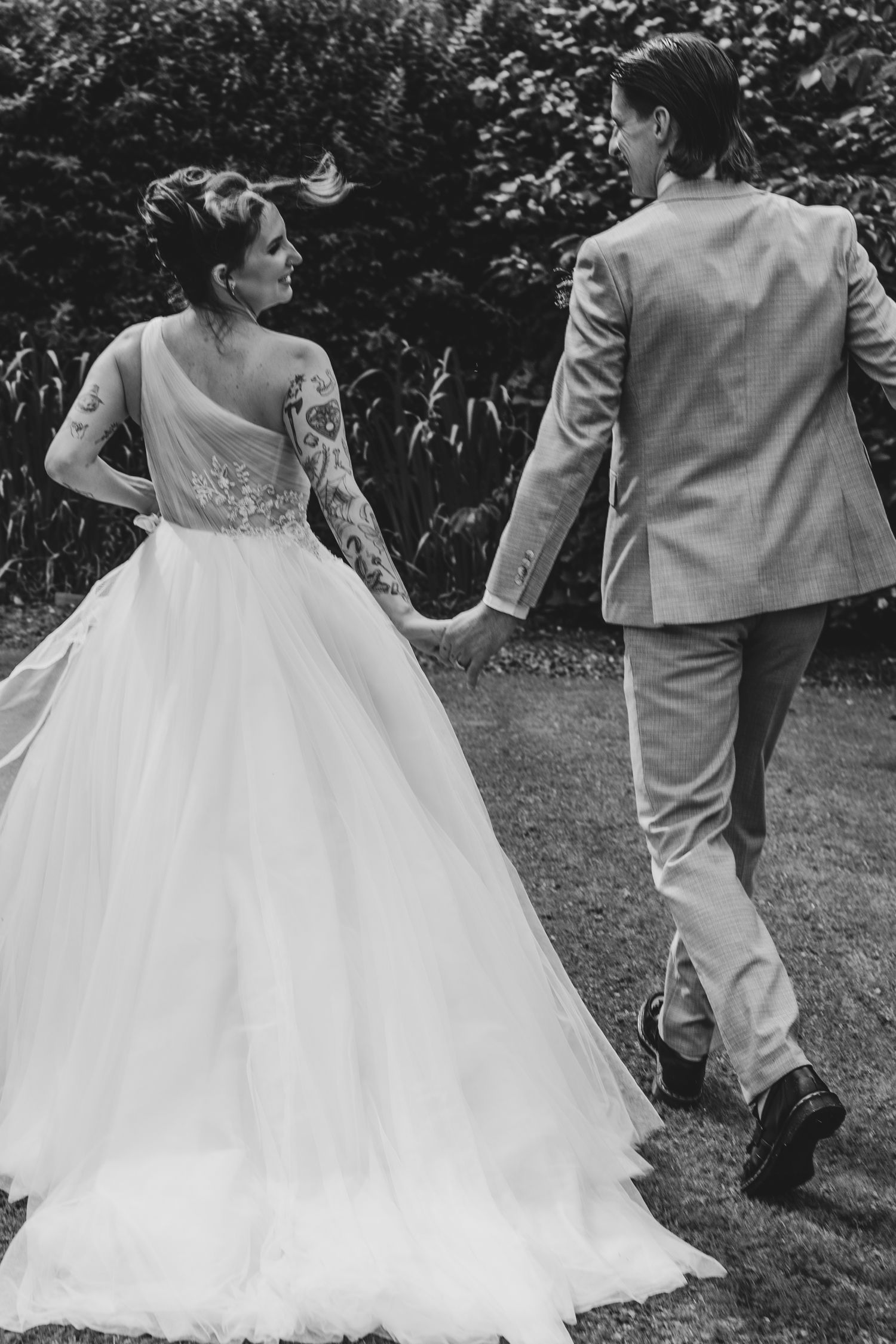 Black and white photo of a tattooed bride in a ballgown holding hands with a suited groom walking through a garden.