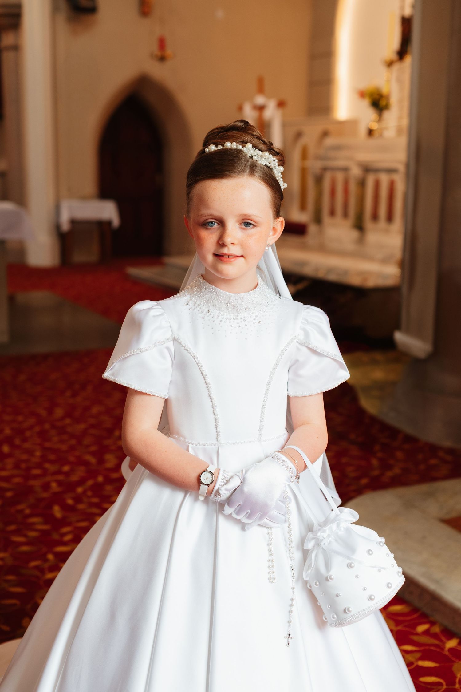 Young girl in white First Communion dress, veil, gloves, and pearl purse smiling inside a church.
