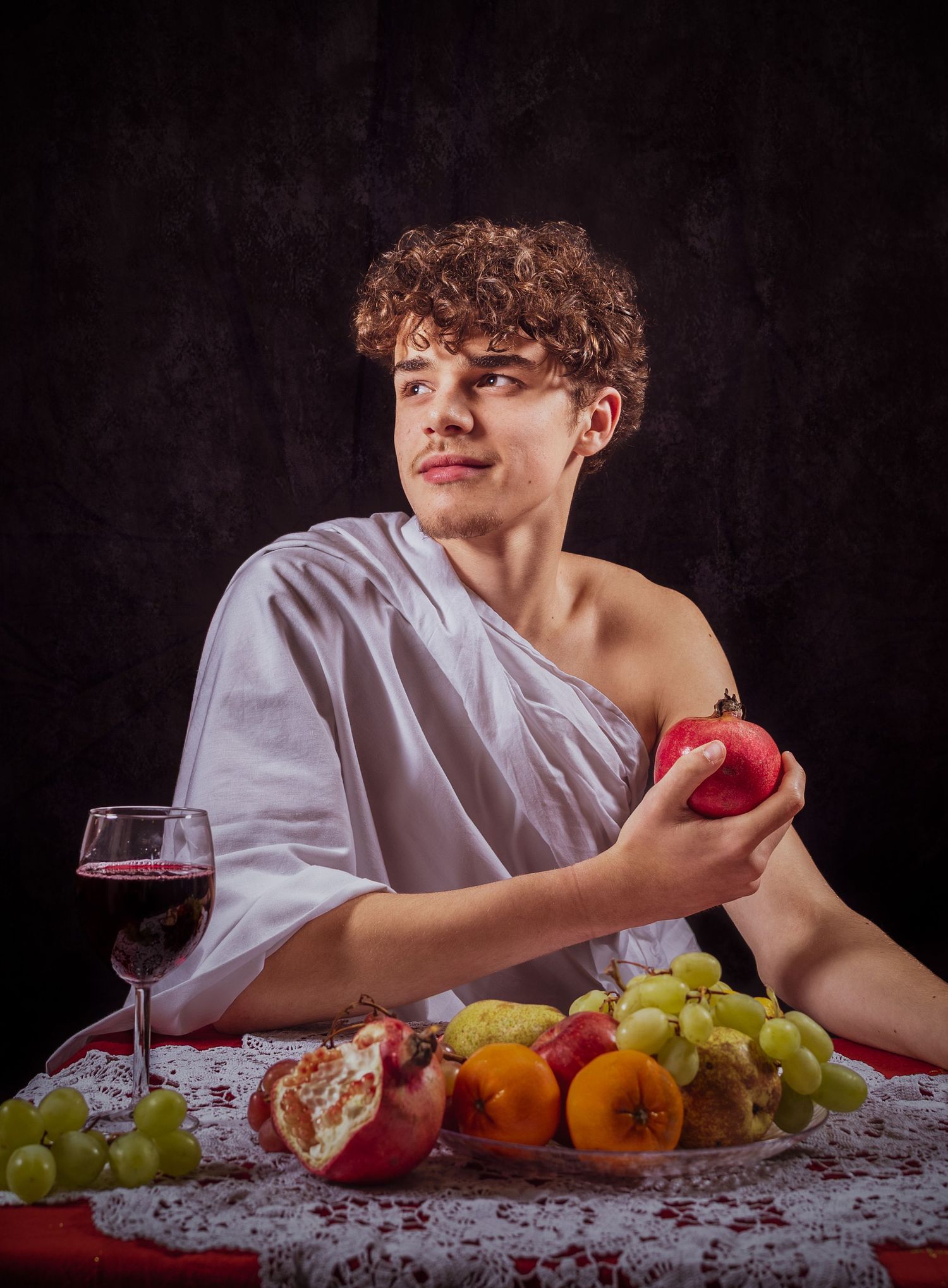 Young man in Roman toga holds pomegranate beside fruit platter and red wine glass on lace tablecloth.