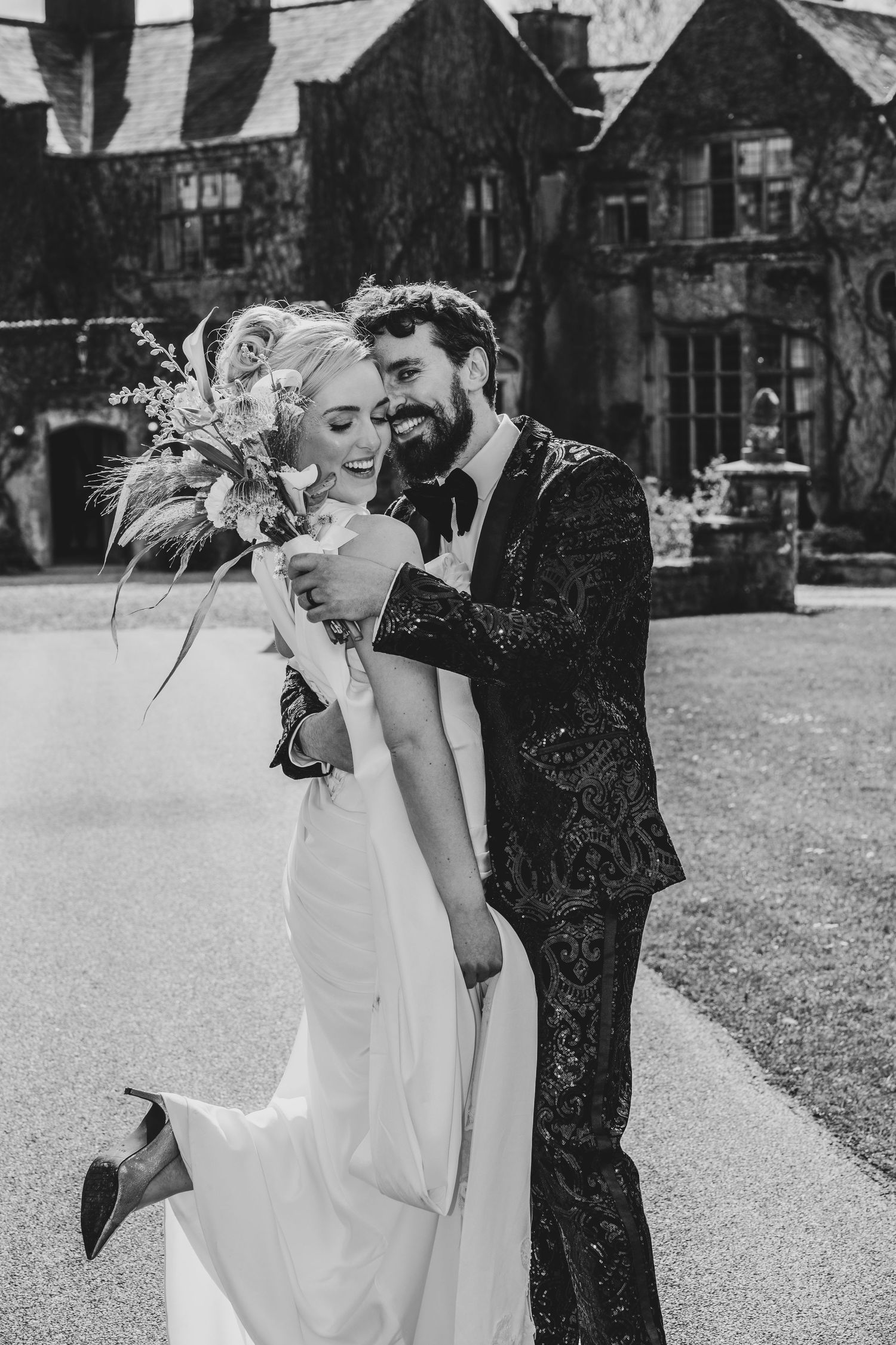 Black and white photo of a smiling bride and groom embracing outdoors at a manor house wedding venue.