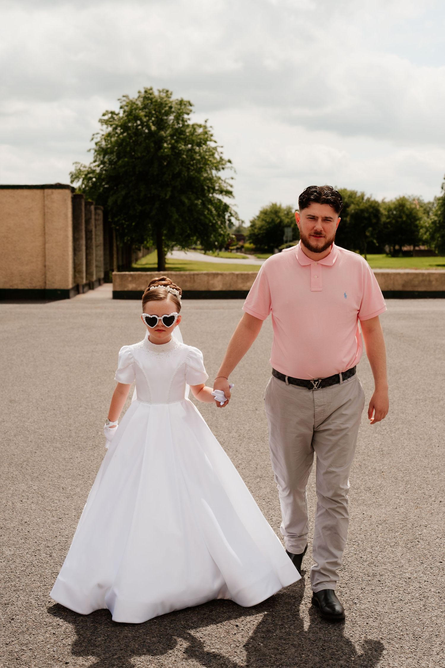 A man in a pink polo holds hands with a young girl in a white communion dress and heart sunglasses outdoors.