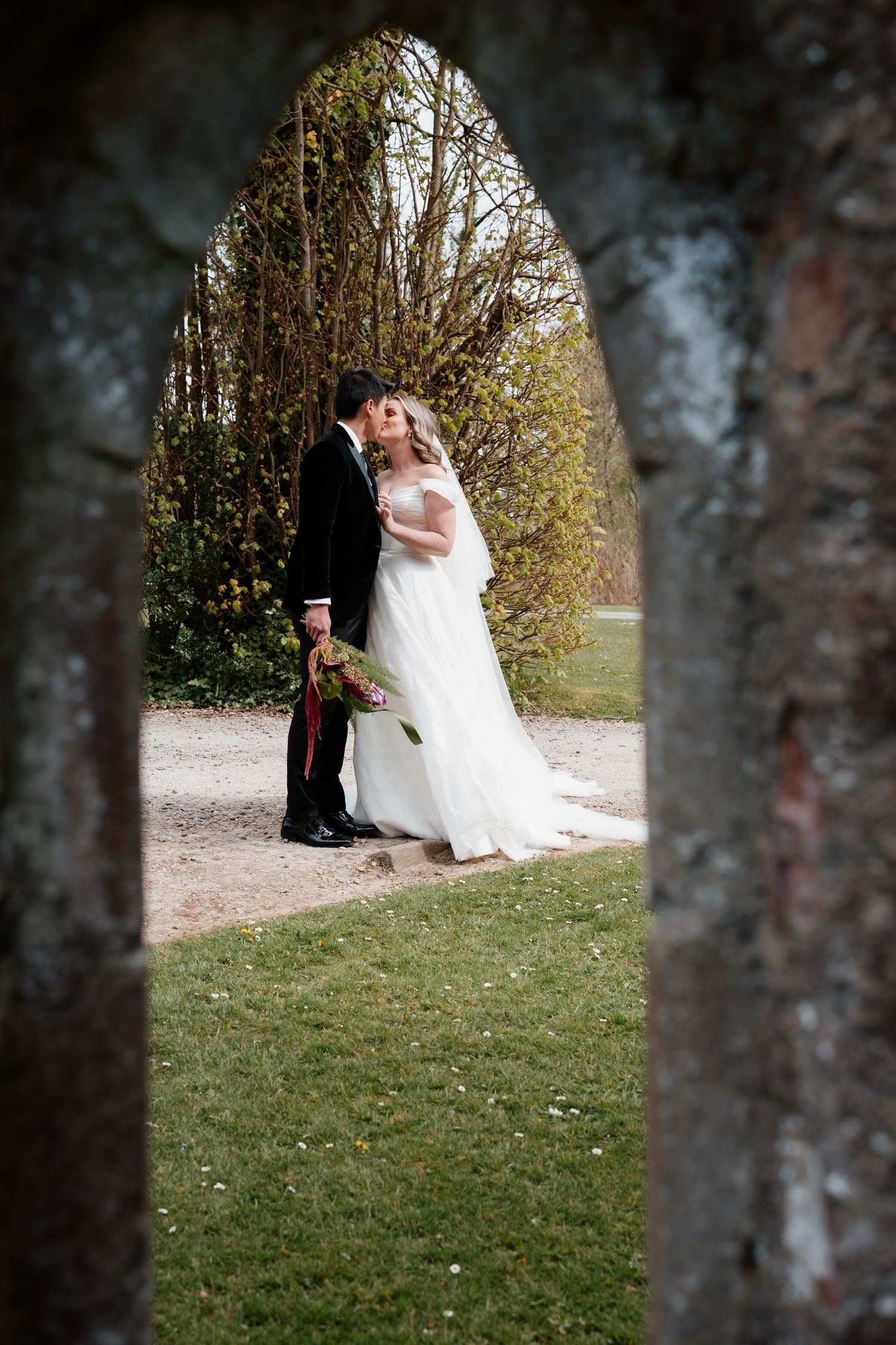 Bride and groom sharing a romantic moment framed by a stone arch in a lush garden setting.