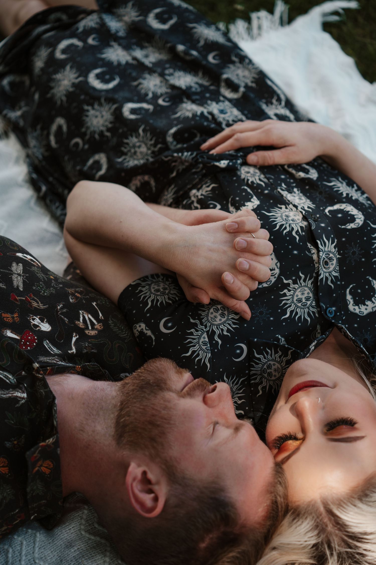 Couple lying on white blanket holding hands, wearing matching celestial print outfits, sharing an intimate moment outdoors.