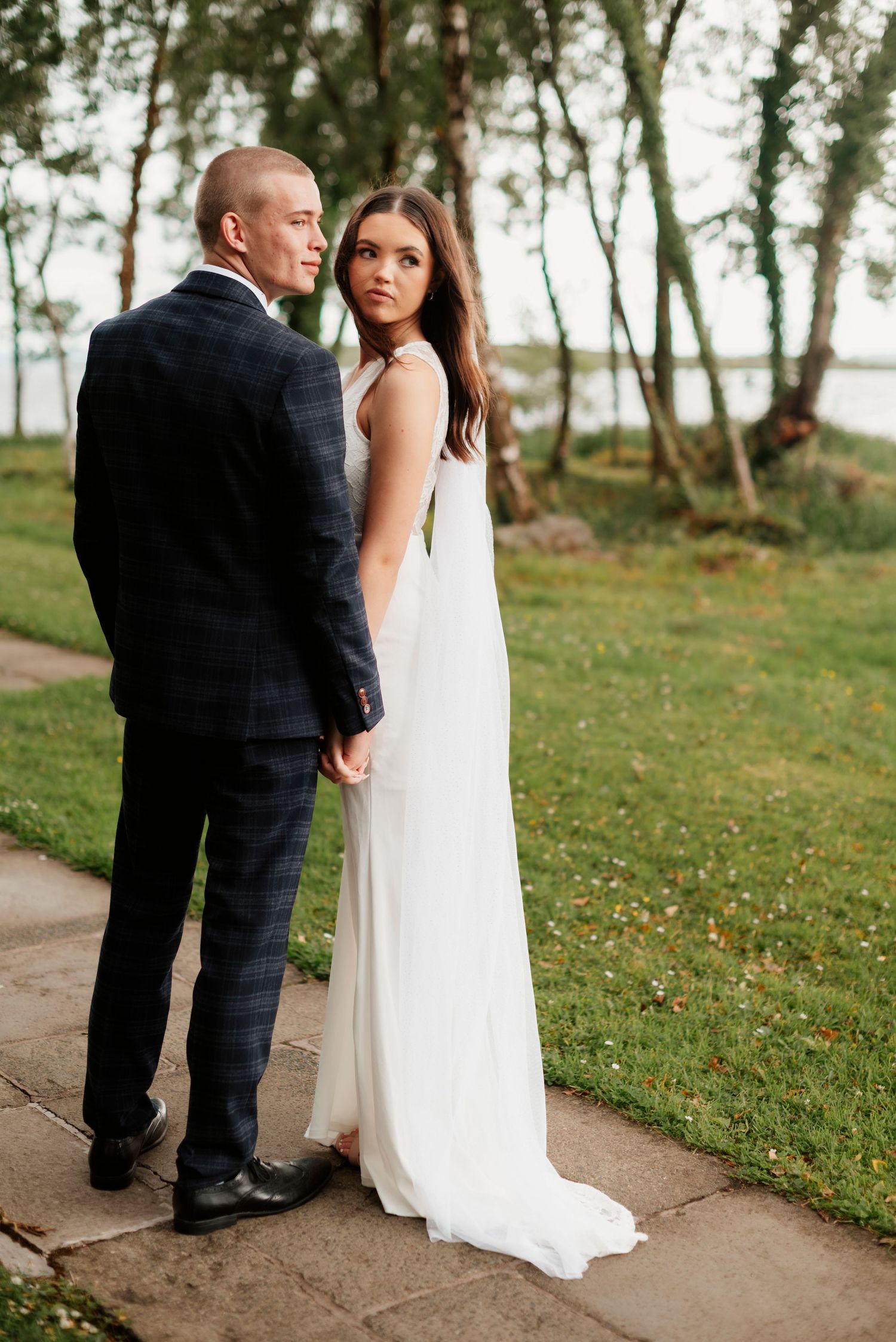 Bride in white gown and groom in plaid suit hold hands on garden path near lake.
