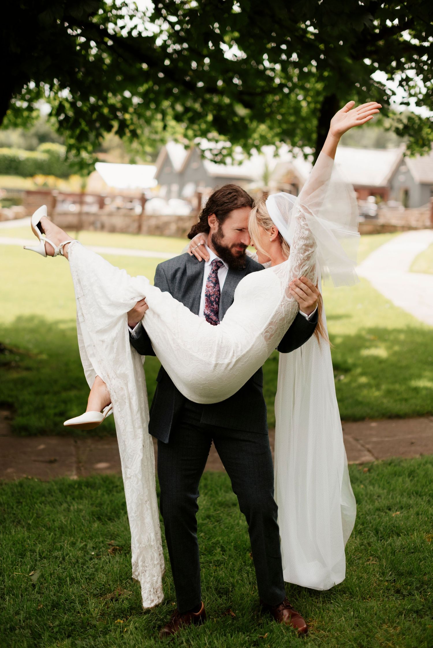 Groom in grey suit lifting bride in white lace gown outdoors on a green lawn.