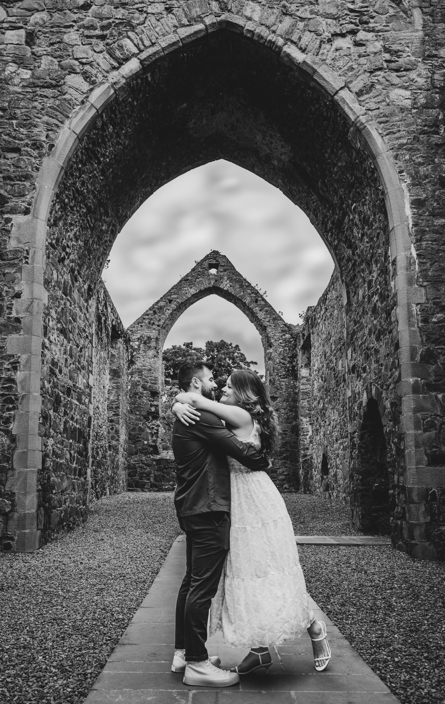Black and white photo of a couple embracing under a Gothic stone arch in ancient ruins.