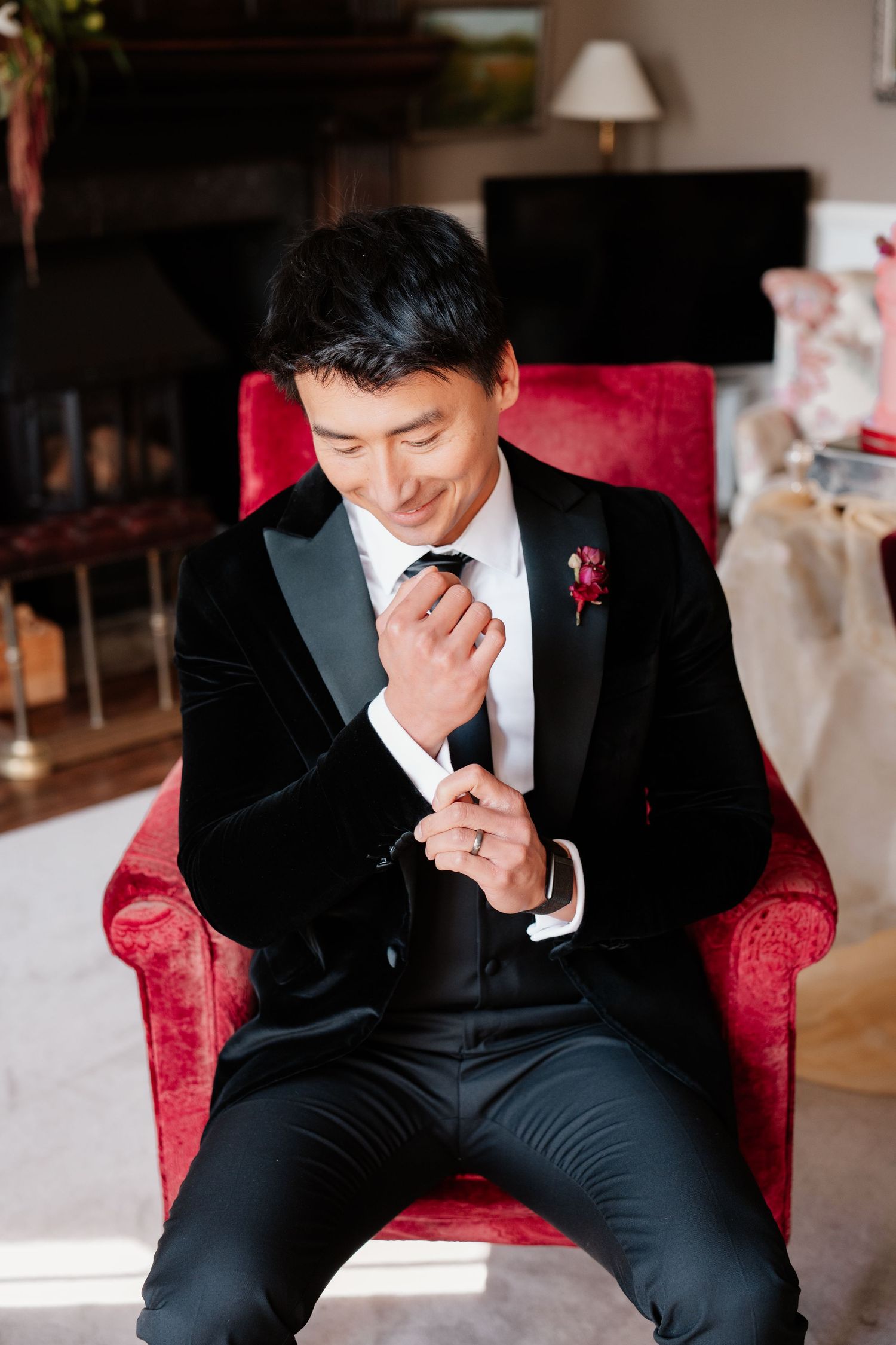Dapper groom in black velvet tuxedo with red boutonniere seated in red velvet chair adjusting cufflinks.