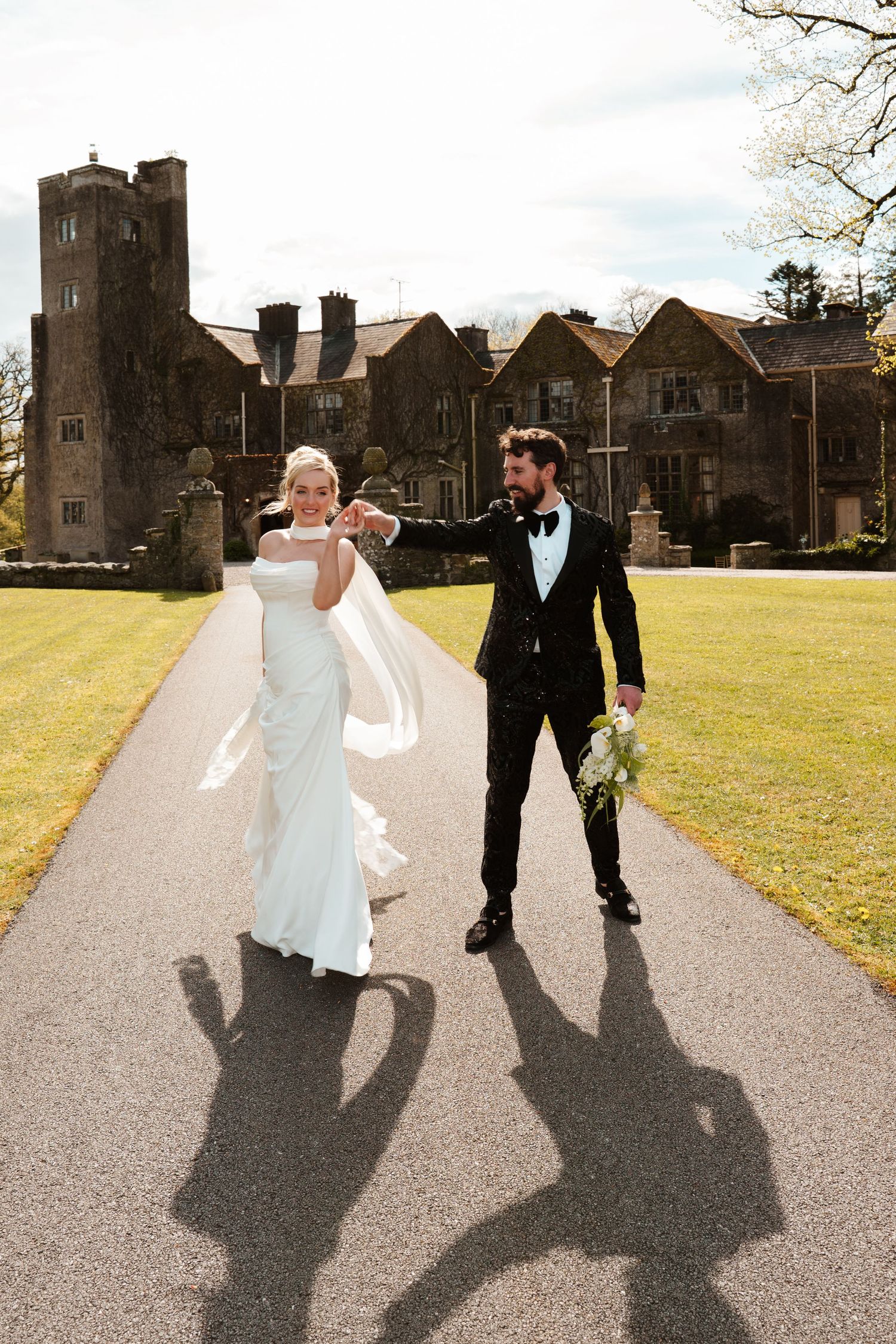 Bride and groom walk hand-in-hand on sunlit path before grand manor house on wedding day.
