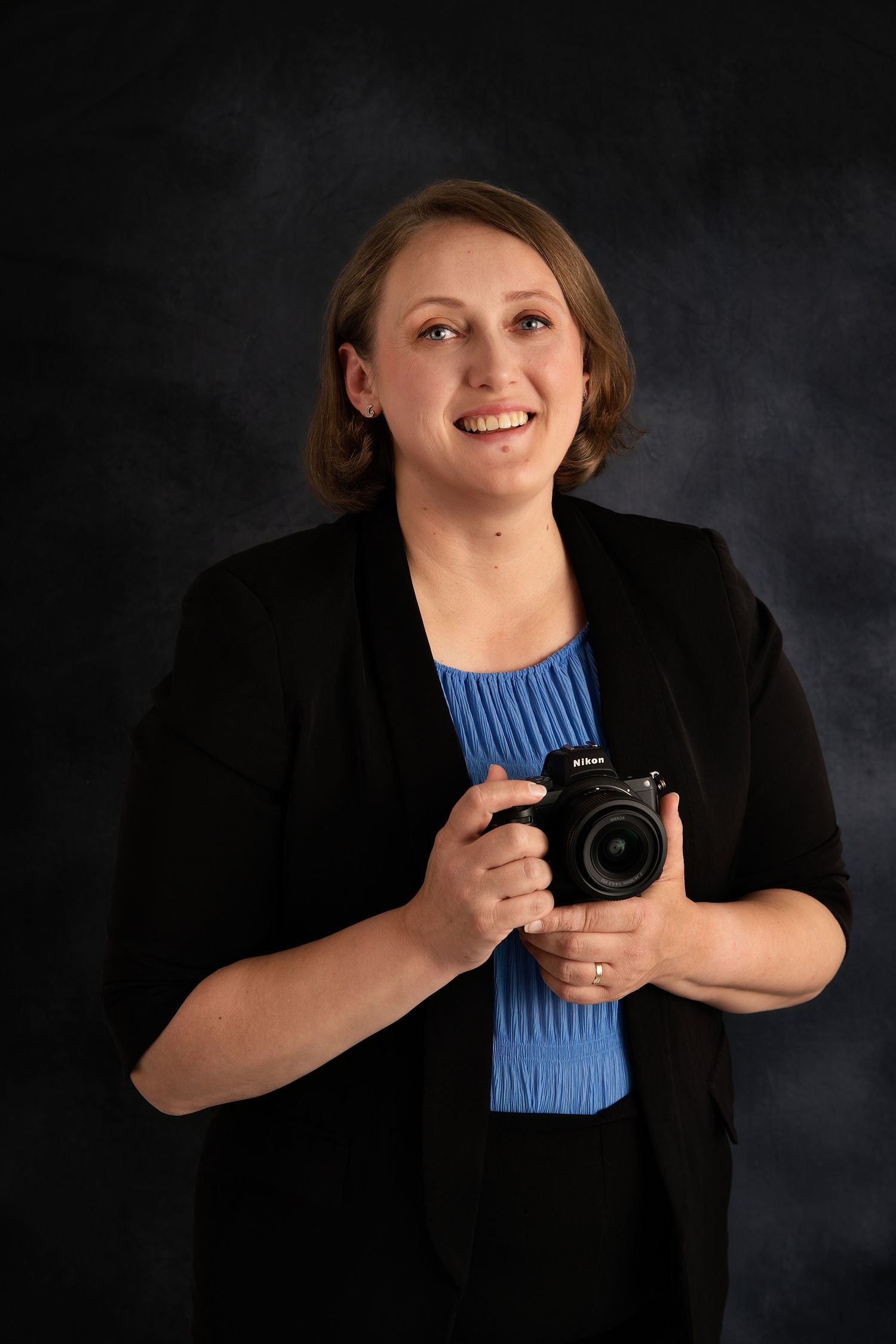 Professional female photographer smiling and holding Nikon camera in studio portrait.