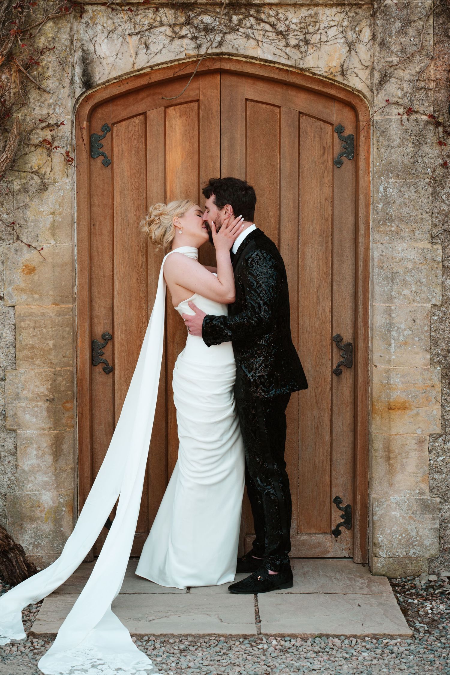 Bride and groom share a romantic kiss in front of a grand wooden arched door, bride wearing a white gown with long cape.