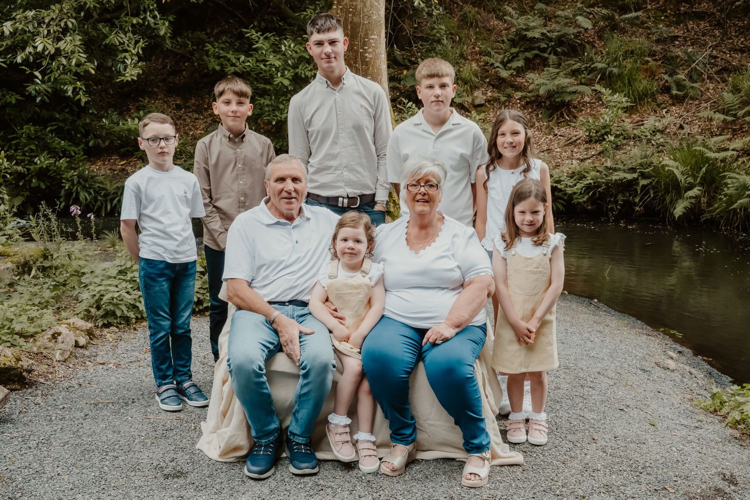 Multi-generational family of nine poses outdoors on a path surrounded by lush green trees near a peaceful stream.